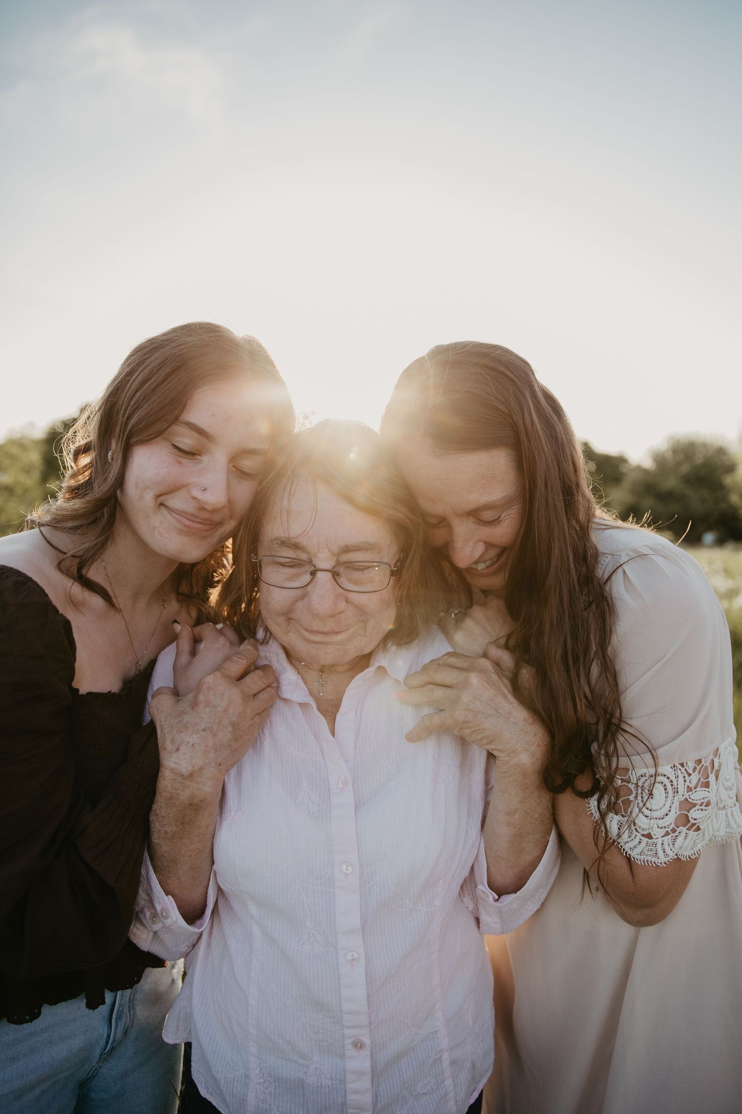 Two young women embrace an older woman outdoors at golden hour, all smiling warmly in a heartfelt family portrait.