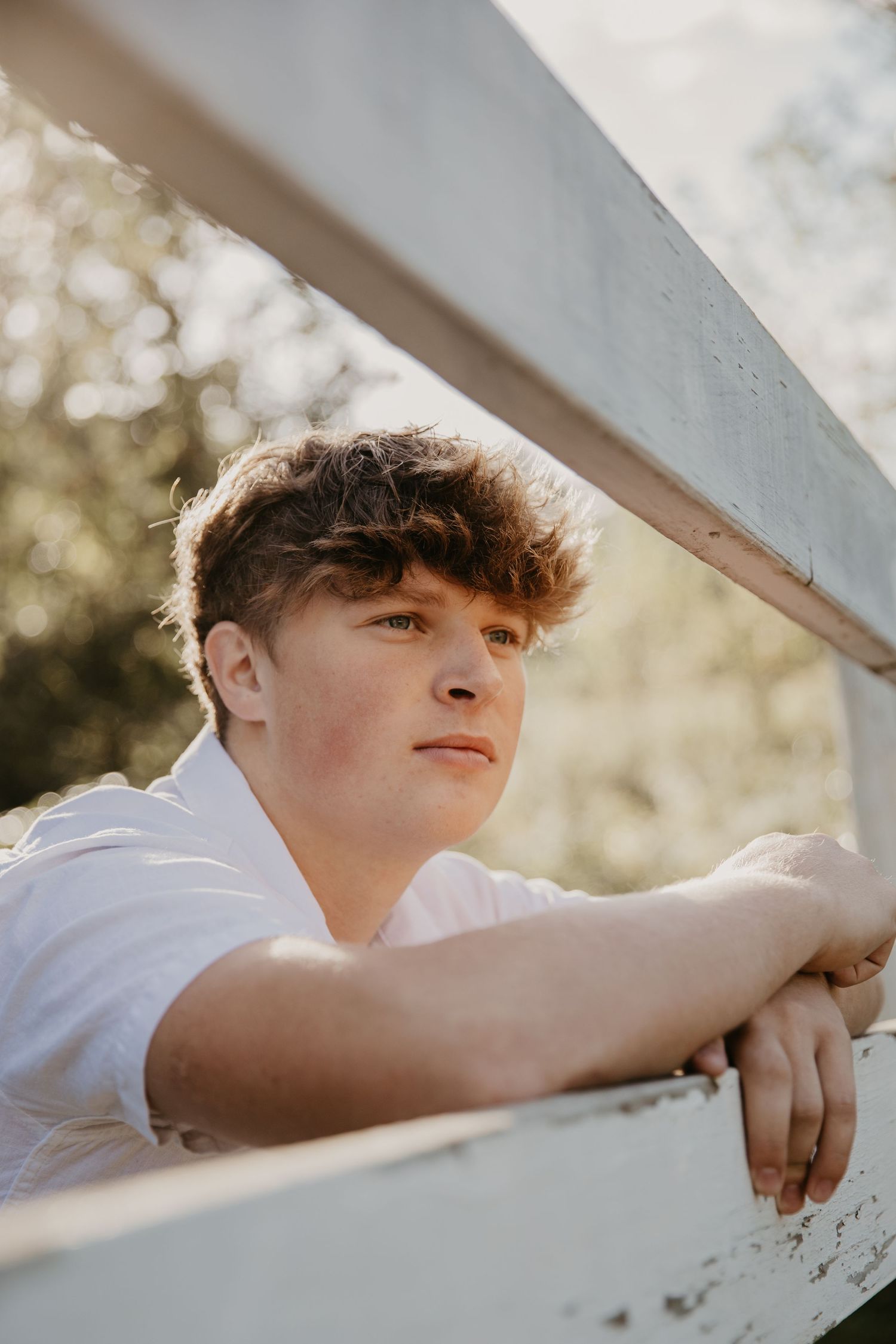 Young man with curly hair leaning on a white wooden fence outdoors, looking thoughtfully into the distance.