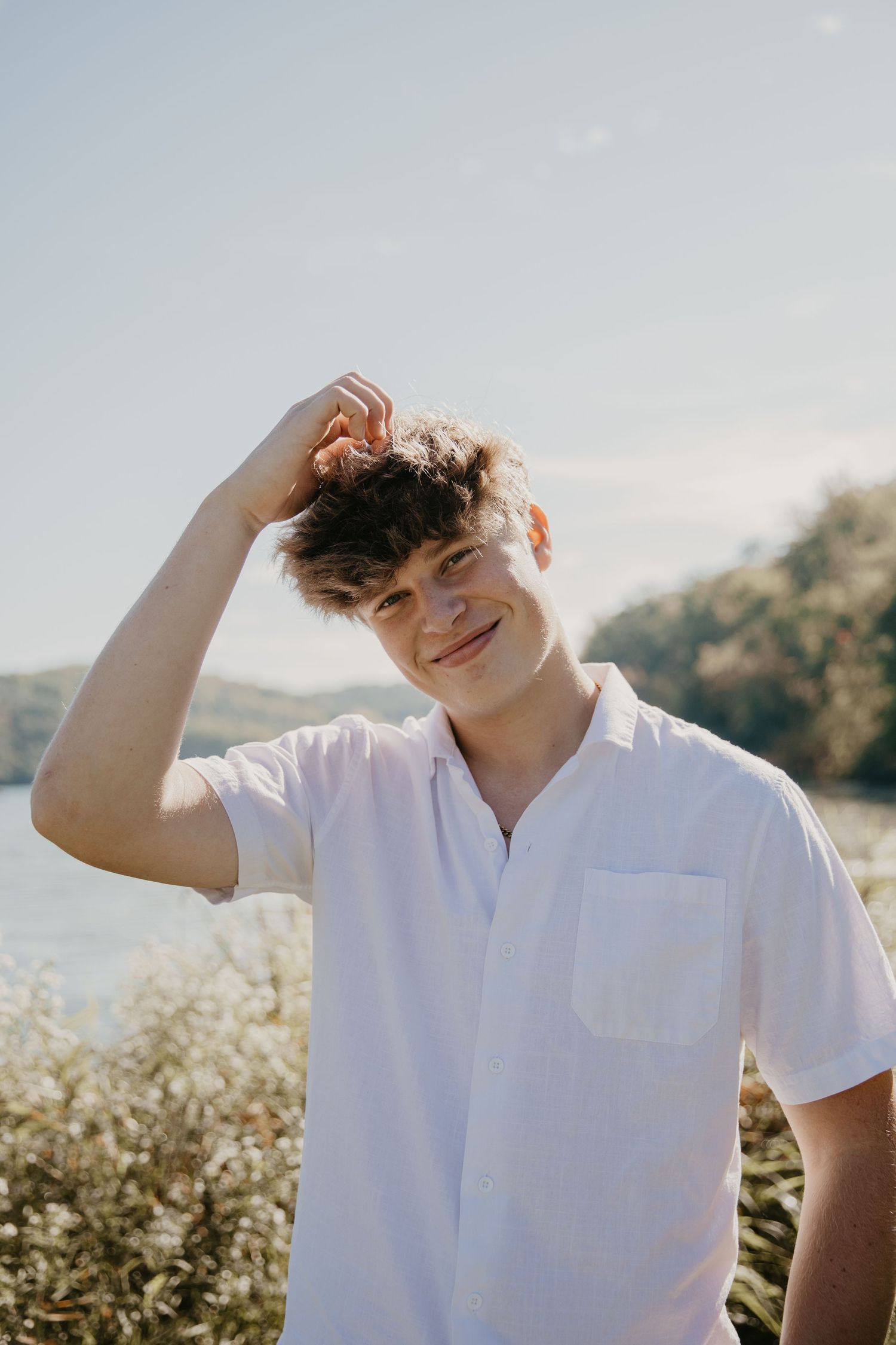 Young man in white polo shirt smiling outdoors near a lake, hand raised to his hair on a sunny day.