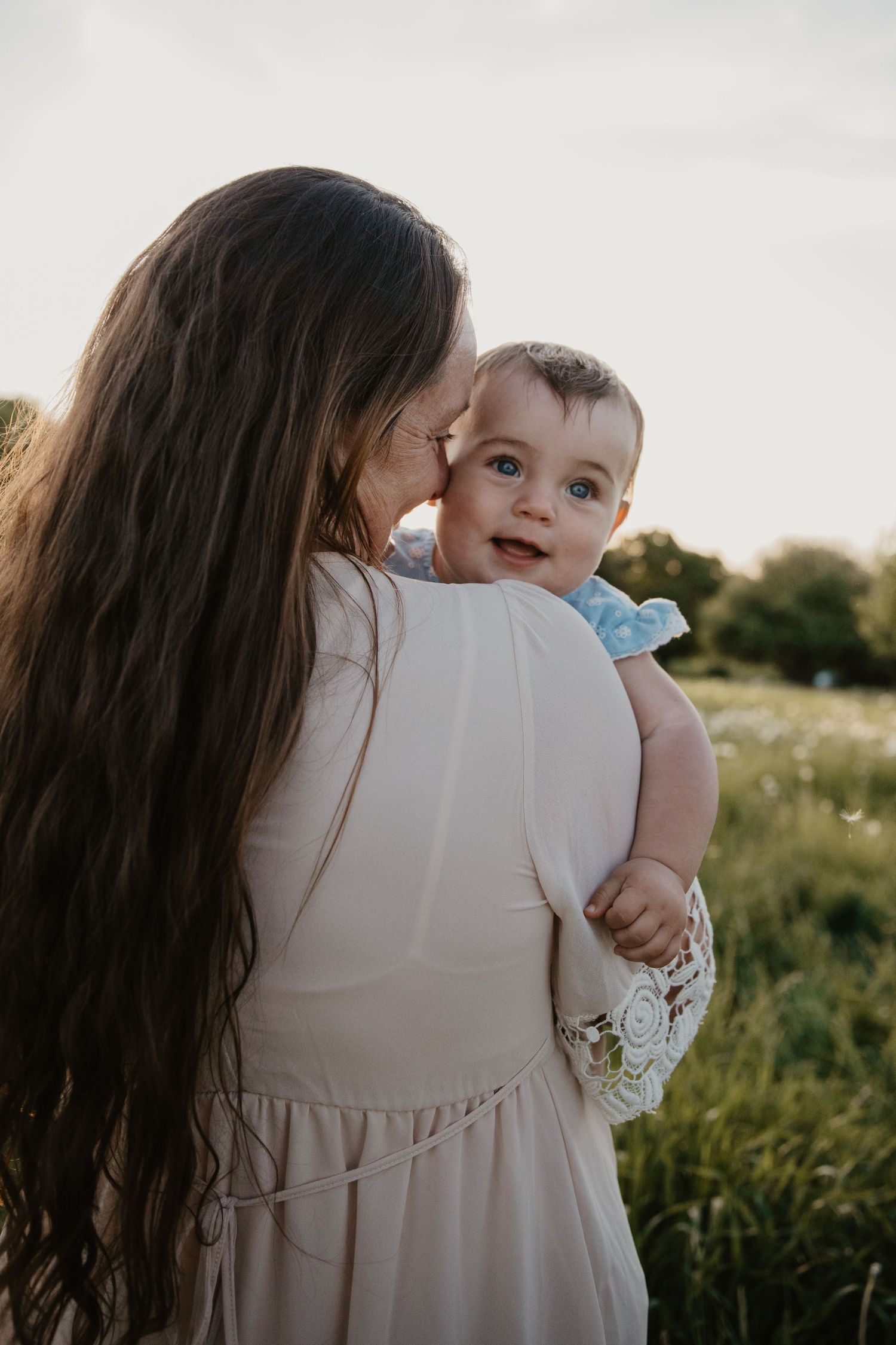 A mother in a white dress kisses her smiling baby outdoors in a sunlit green field at golden hour.