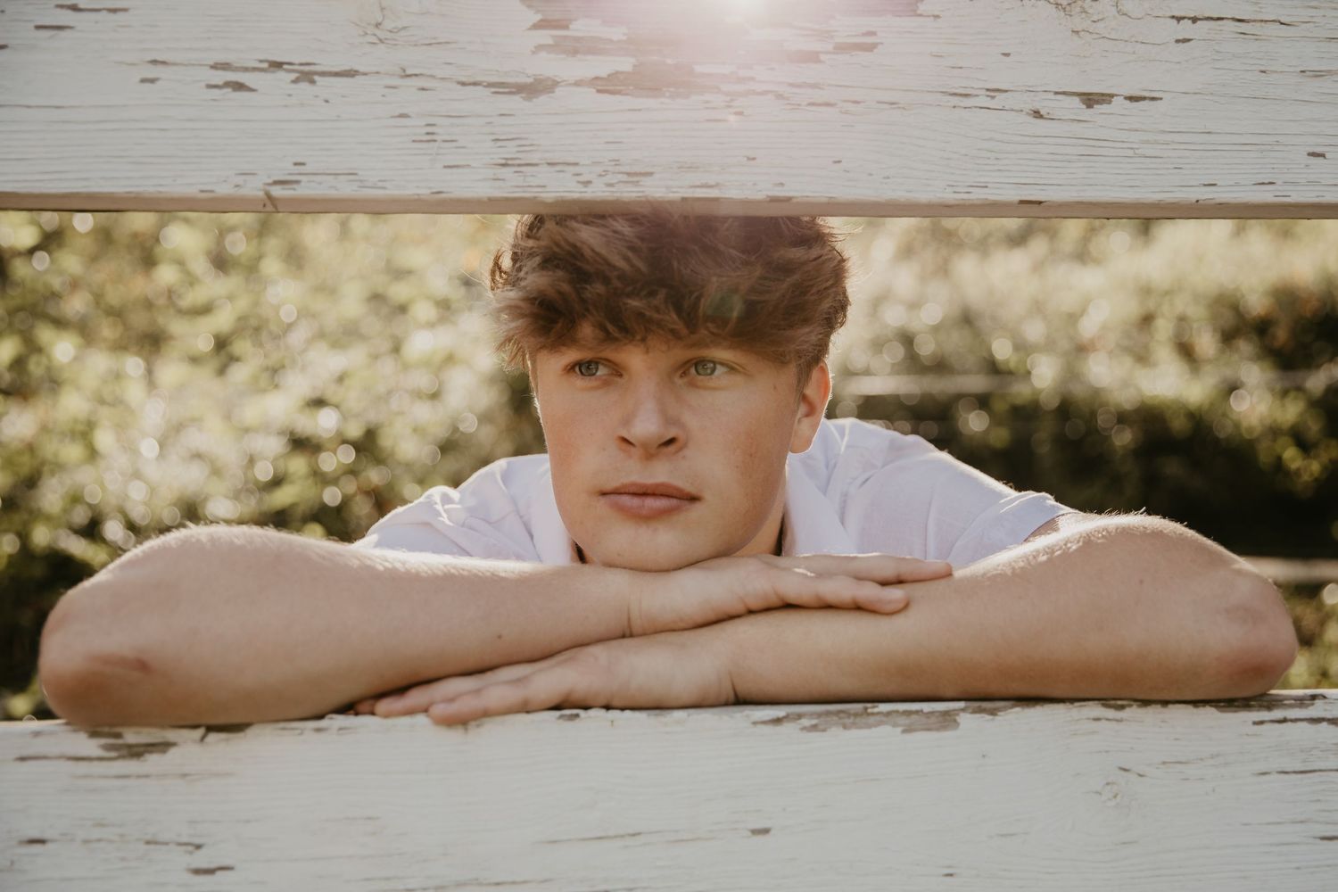 Young man with brown hair resting chin on folded arms on a white wooden surface outdoors.