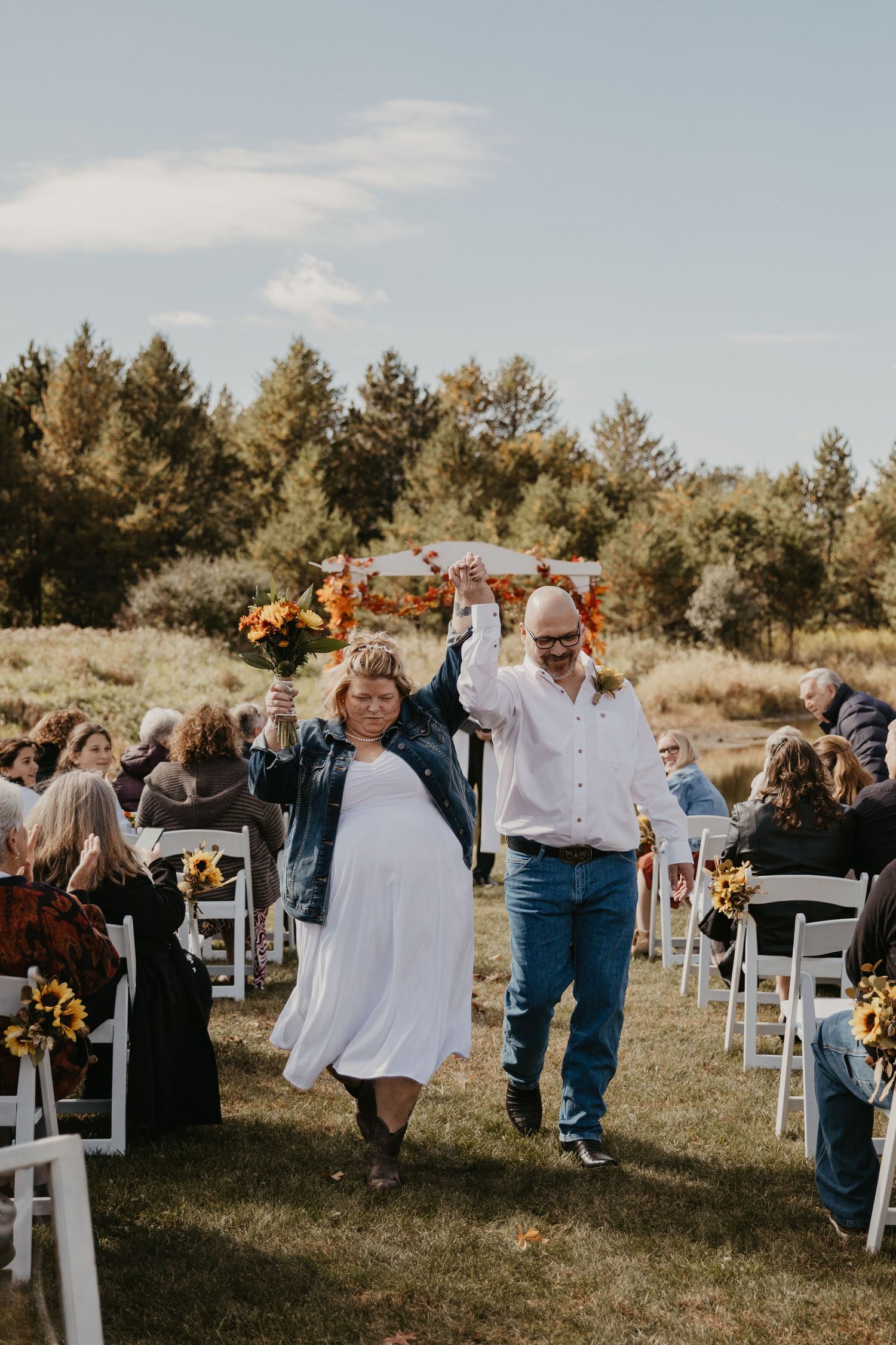 Joyful newlyweds raise hands walking down outdoor fall wedding aisle lined with sunflowers and white chairs.