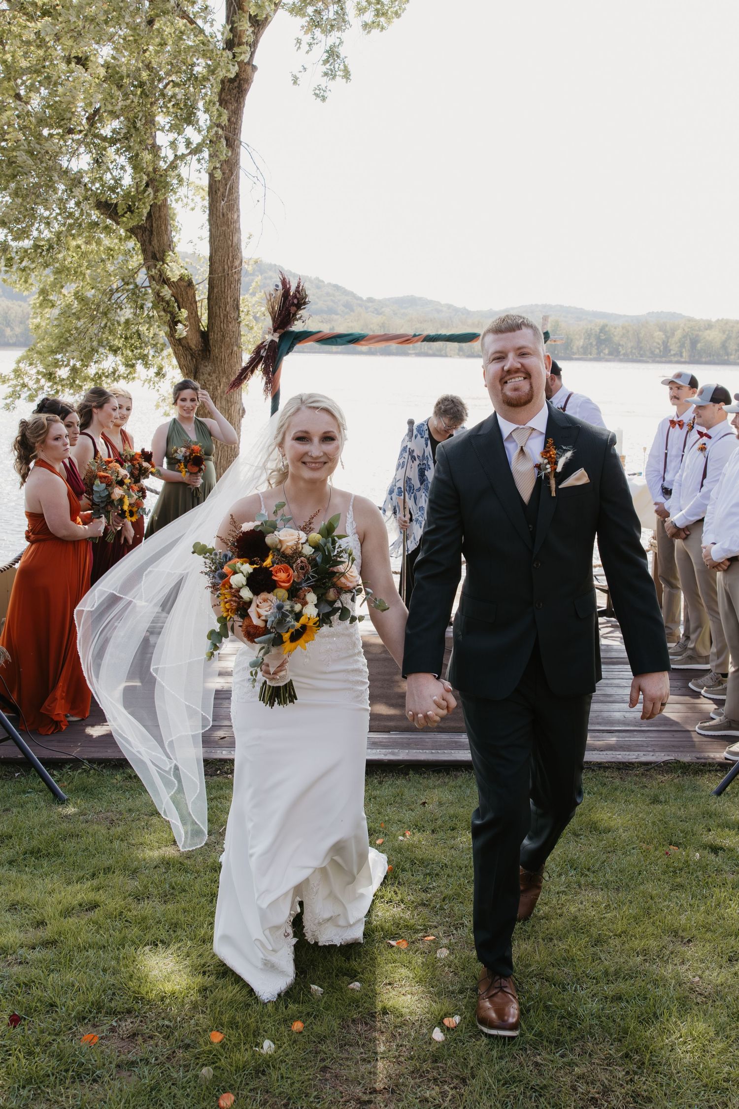 Bride and groom walk hand-in-hand after outdoor lakeside wedding ceremony, bride holding colorful autumn bouquet with veil flowing.