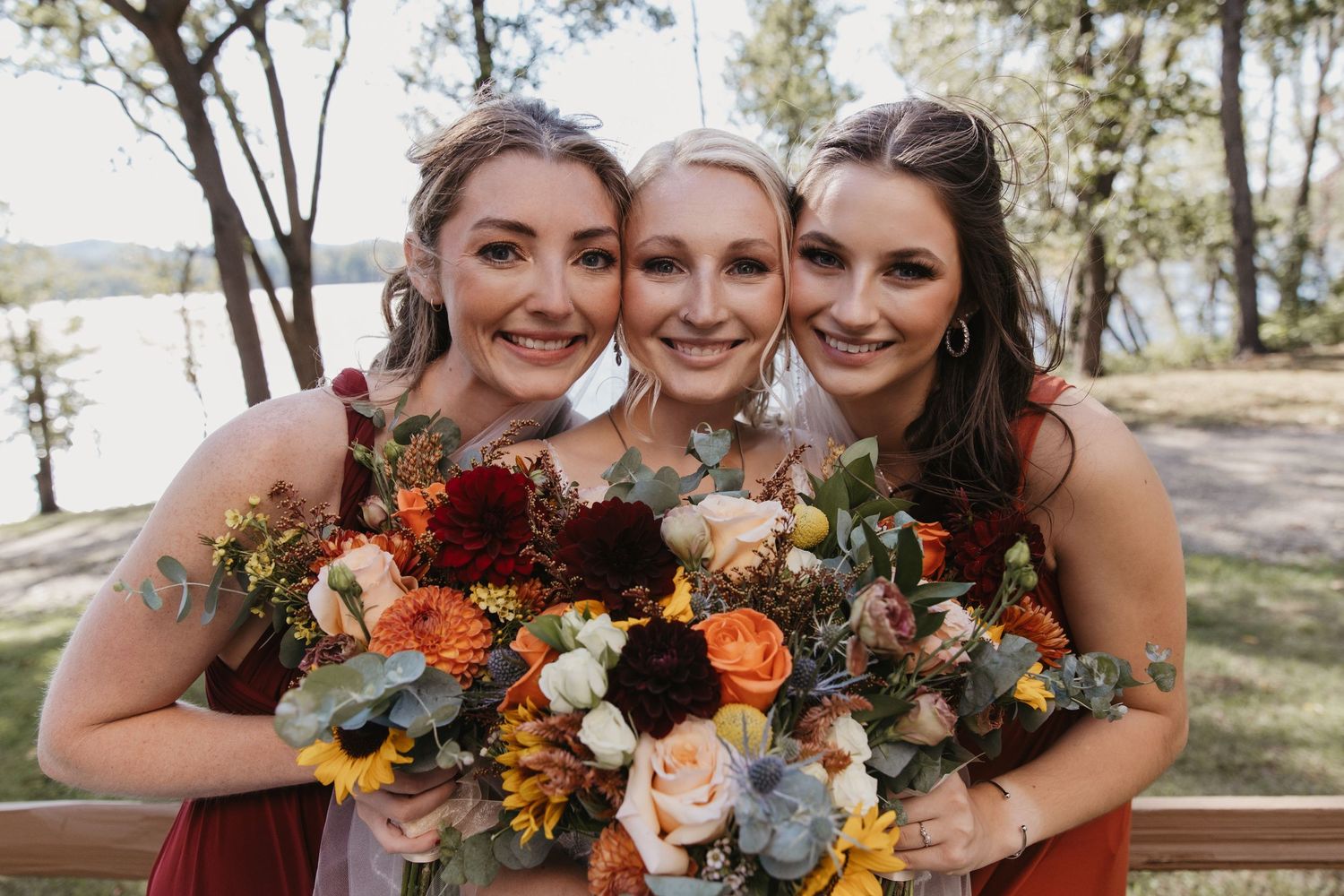 Three bridesmaids smiling together outdoors holding colorful autumn bouquets with red, orange, and white flowers near a lake.