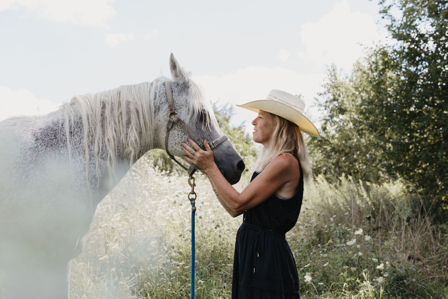 Woman in cowboy hat and black dress tenderly holding a white horse on a blue lead rope outdoors.