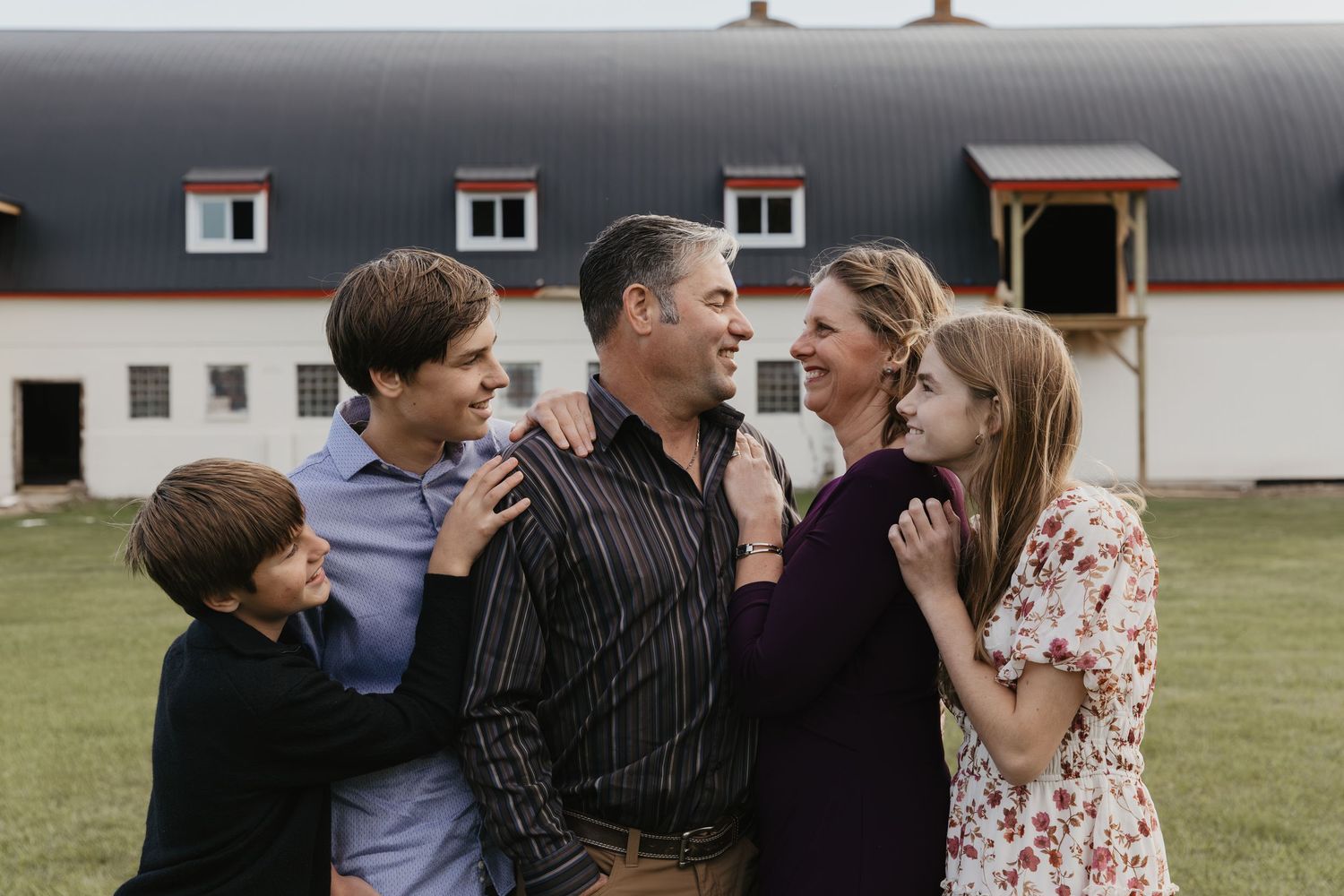 Happy family of five sharing a warm embrace outdoors in front of a white barn farmhouse.