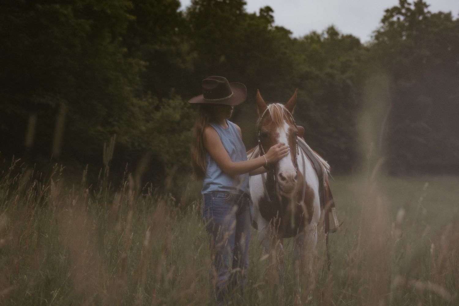A person in a cowboy hat and denim stands in tall grass, gently petting a brown and white horse in a misty, tree-lined meadow.