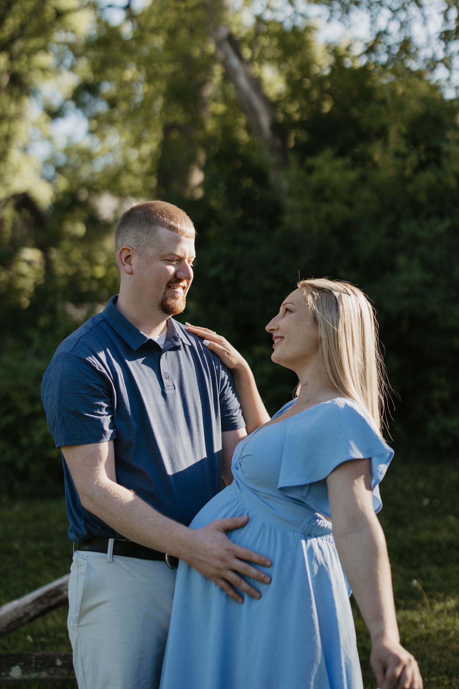 Smiling couple in outdoor maternity photo, man in navy polo holds pregnant womans belly, woman in light blue dress.