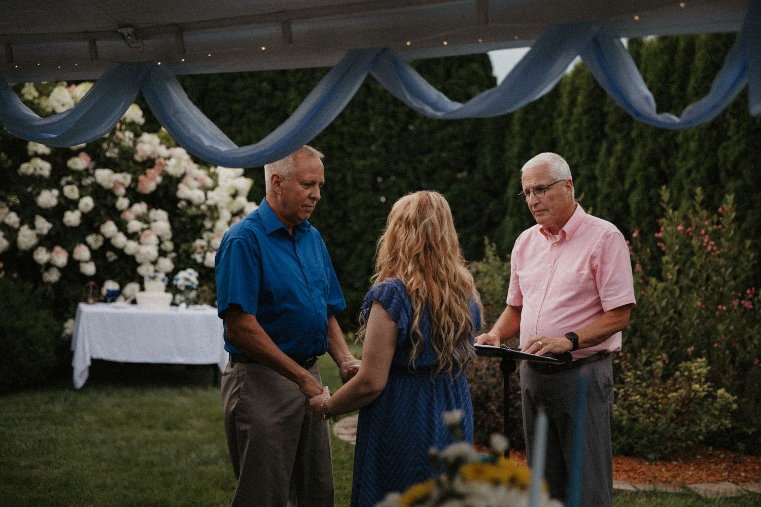 Outdoor wedding ceremony with couple holding hands before officiant, floral arrangements and blue draping in background.