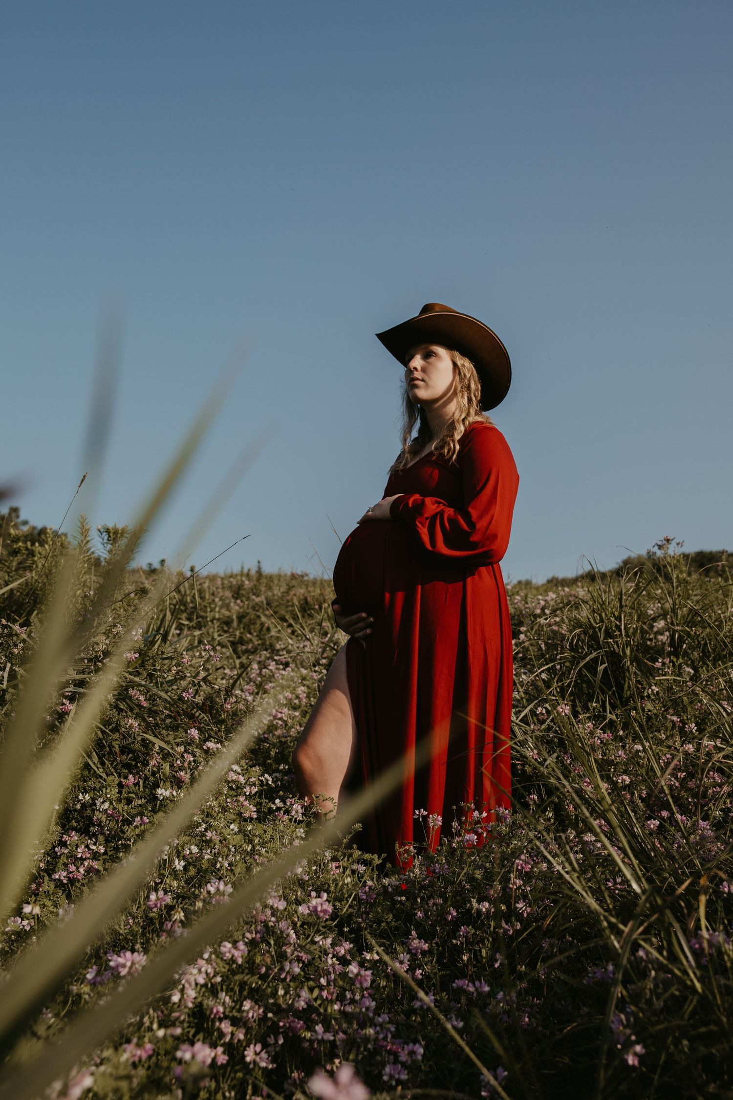 Pregnant woman in red dress and cowboy hat standing in wildflower field under clear blue sky.