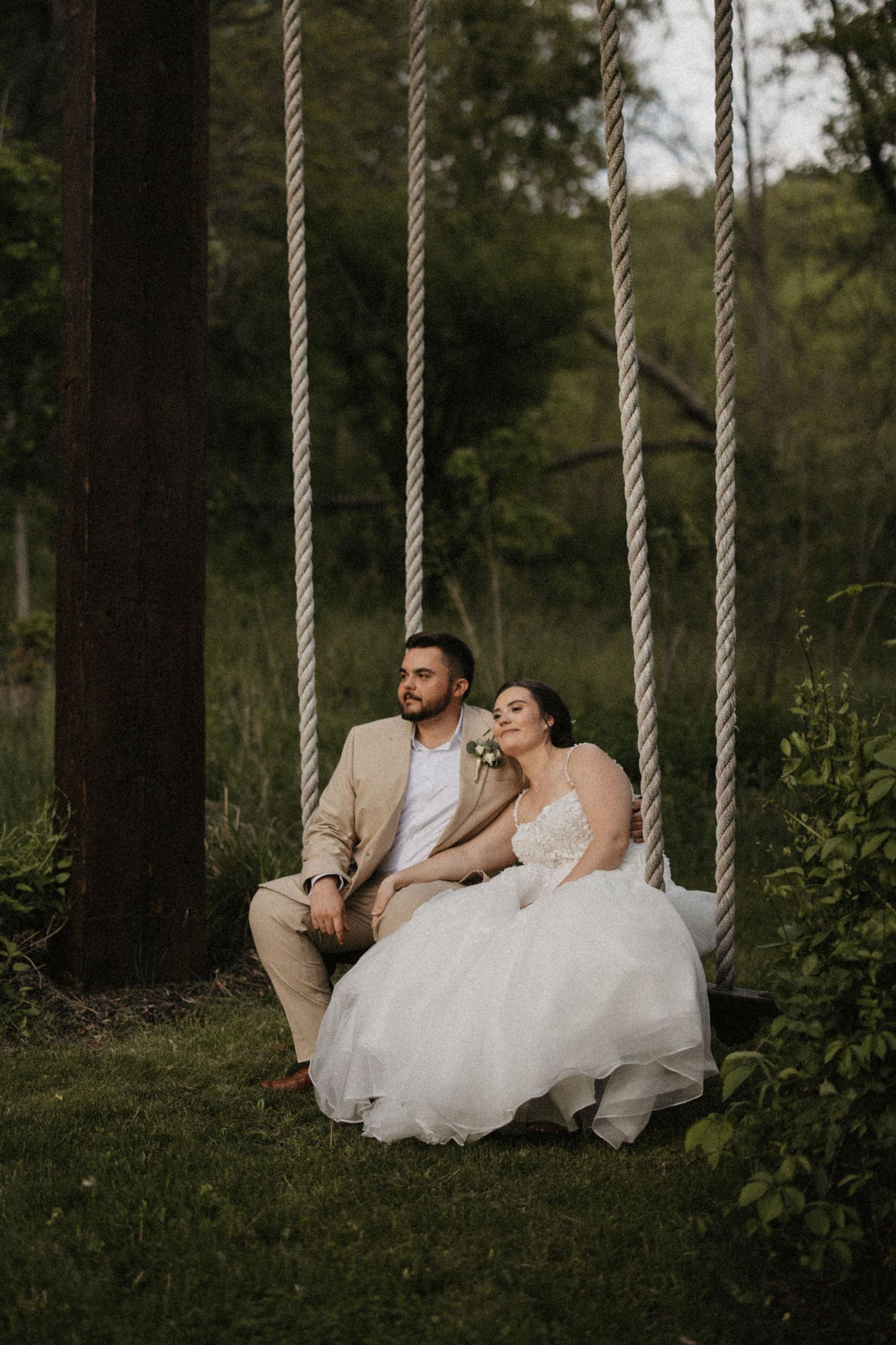 Bride and groom sitting together on a rope swing surrounded by lush green trees in an outdoor wedding setting.