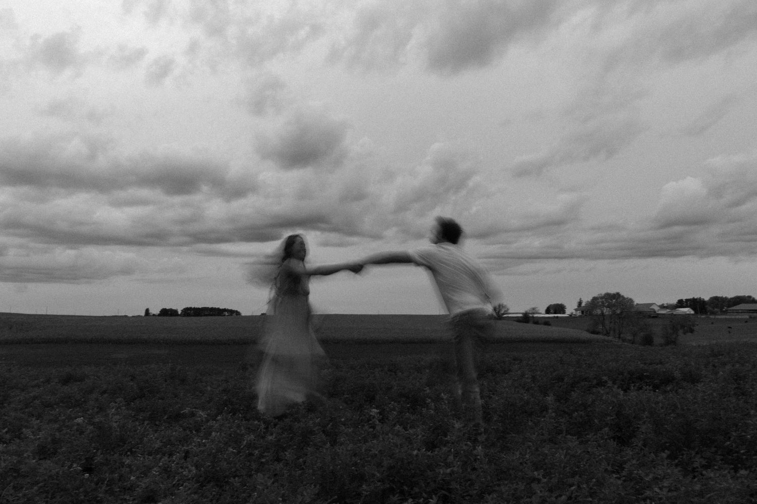 Black and white photo of couple holding hands spinning in field under dramatic cloudy sky.