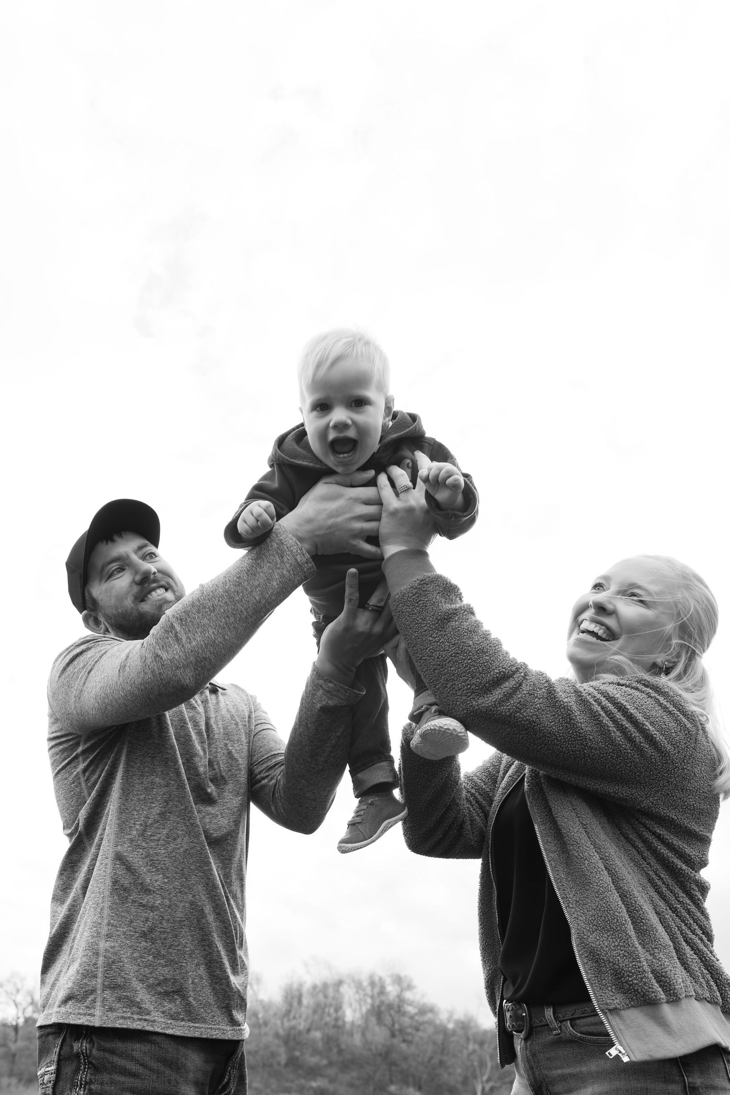 Black and white photo of happy parents lifting their smiling toddler up in the air outdoors.