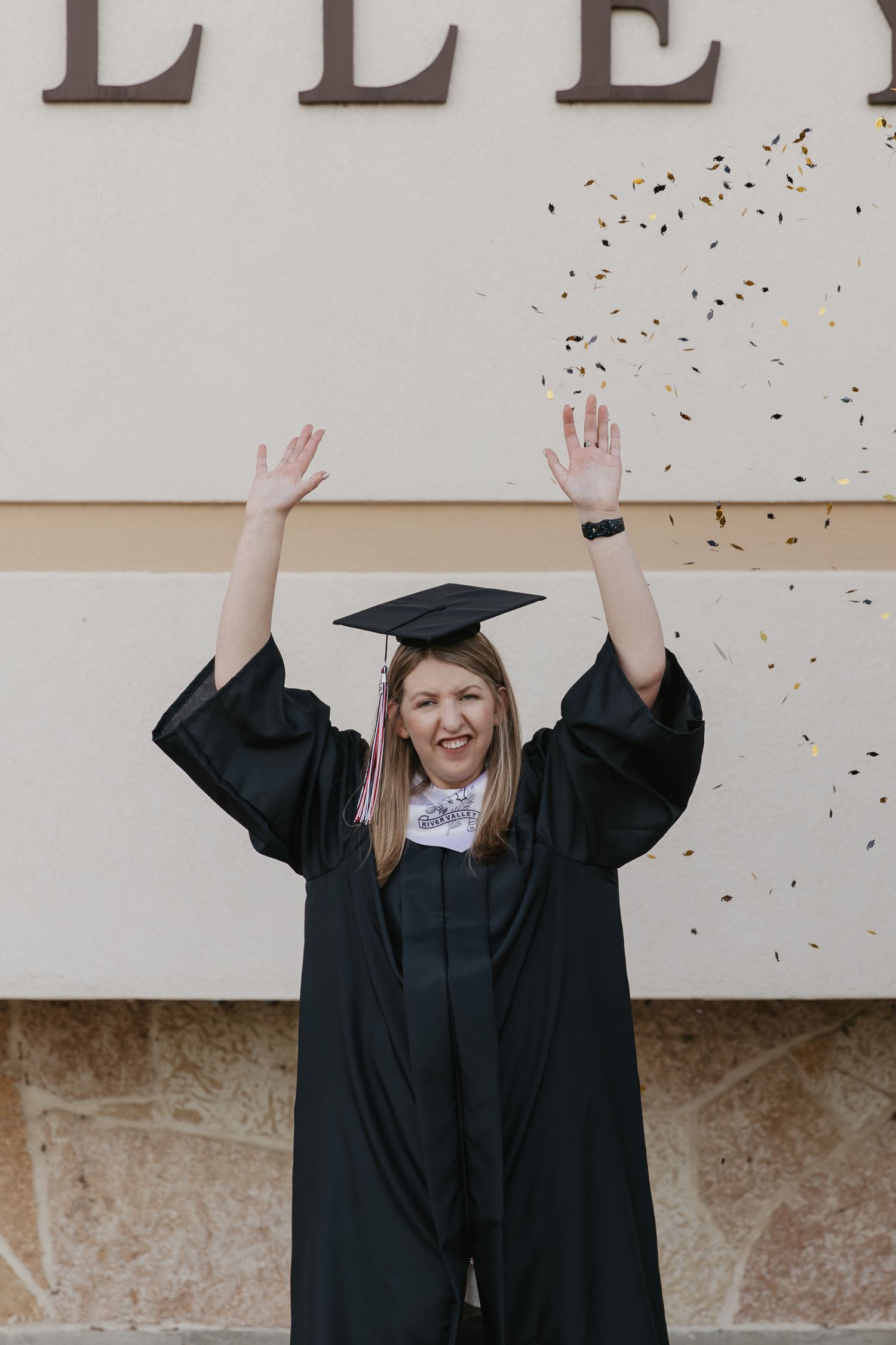 A joyful graduate in black cap and gown raises her arms as gold confetti falls around her outdoors.