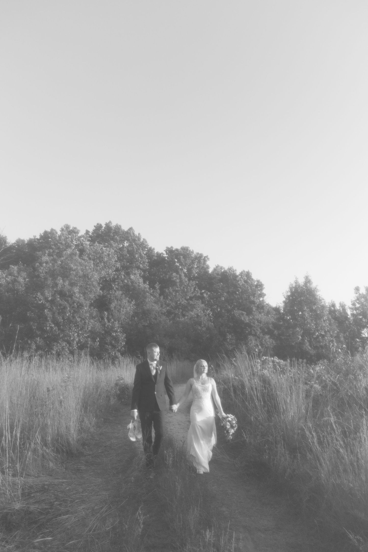 Black and white photo of bride and groom walking hand in hand through a meadow path surrounded by tall grass.