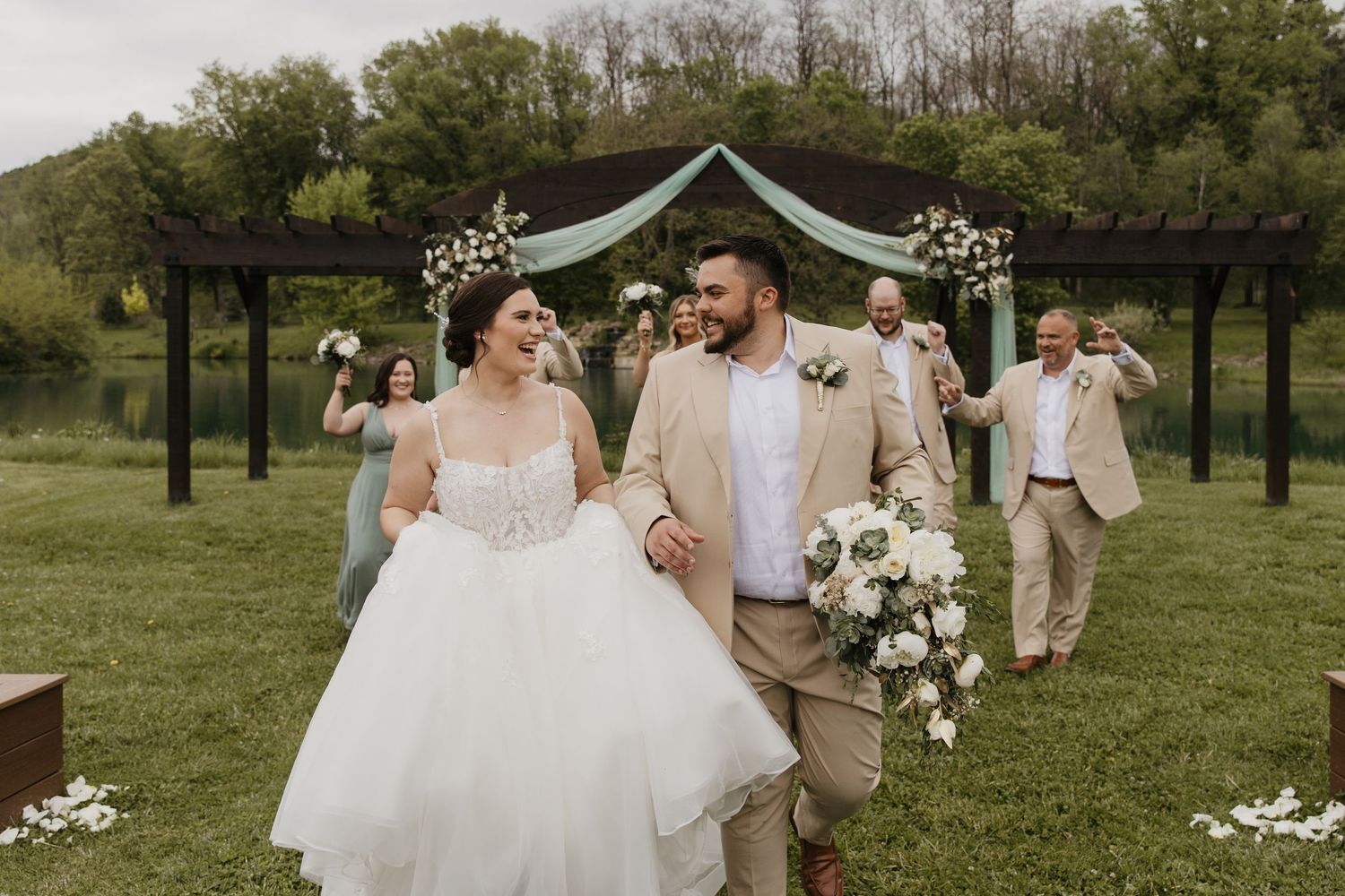 Joyful newlyweds walk down the aisle at an outdoor wedding ceremony with green hills and a floral arch backdrop.