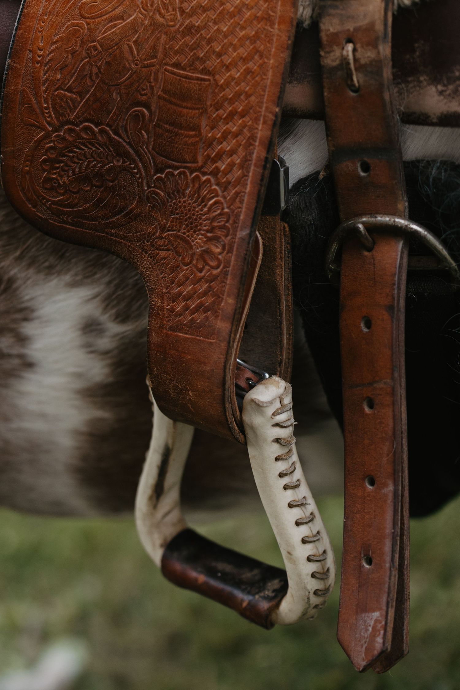 Close-up of a ornate tooled leather western saddle with decorative floral carving, stirrup, and cinch strap on a horse.