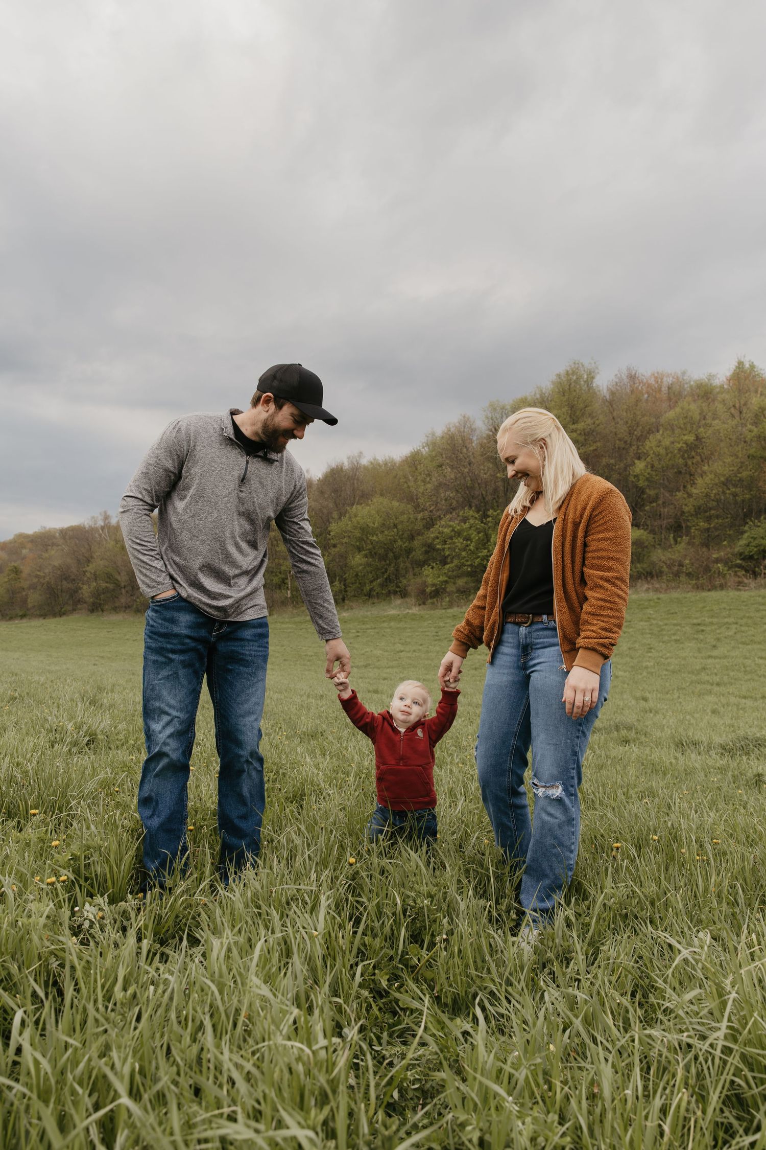 A young family with a toddler holding hands in a green field, with trees and cloudy sky in the background.