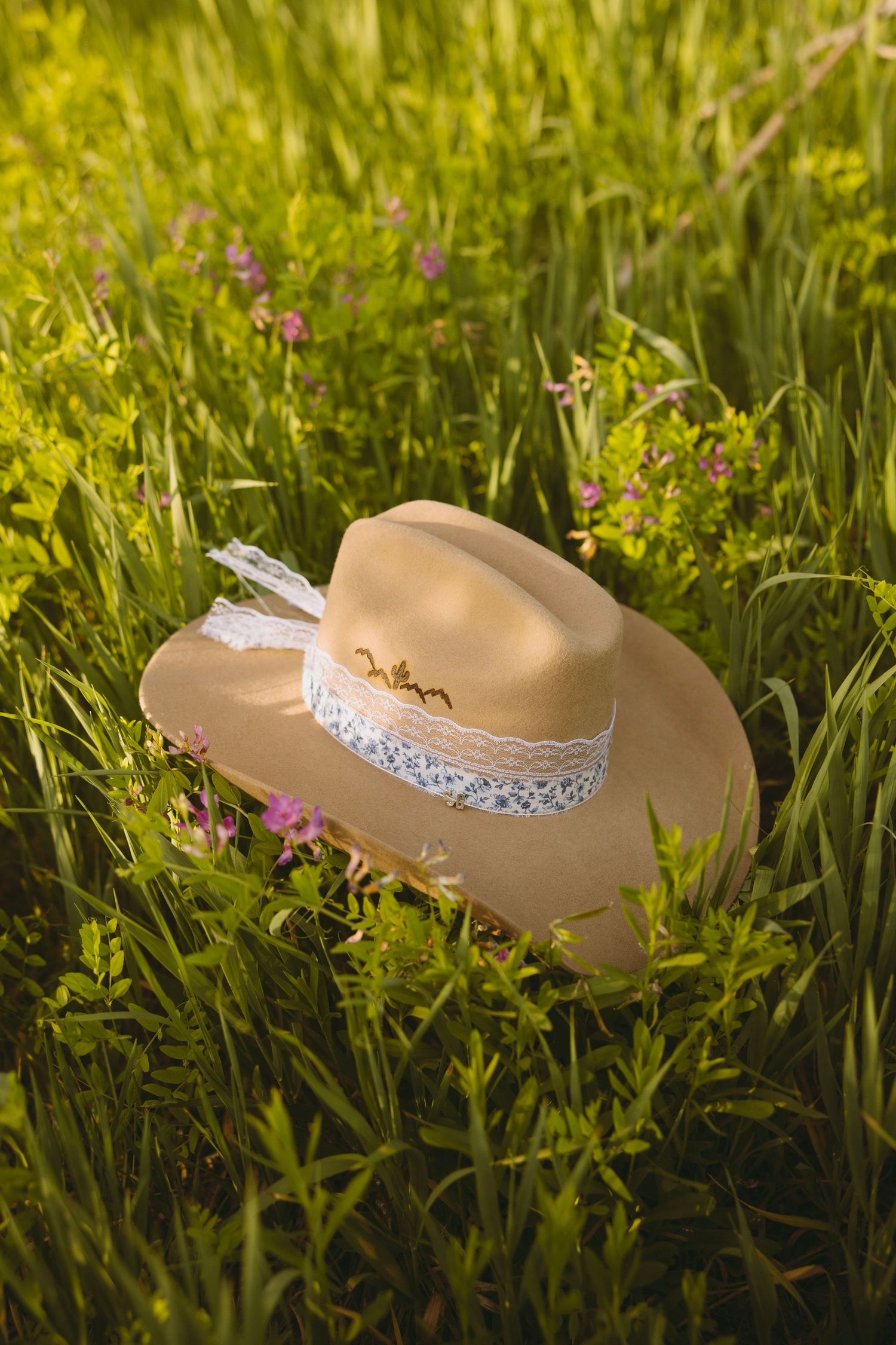A beige sun hat decorated with white lace band rests in green grass among wildflowers in golden sunlight.