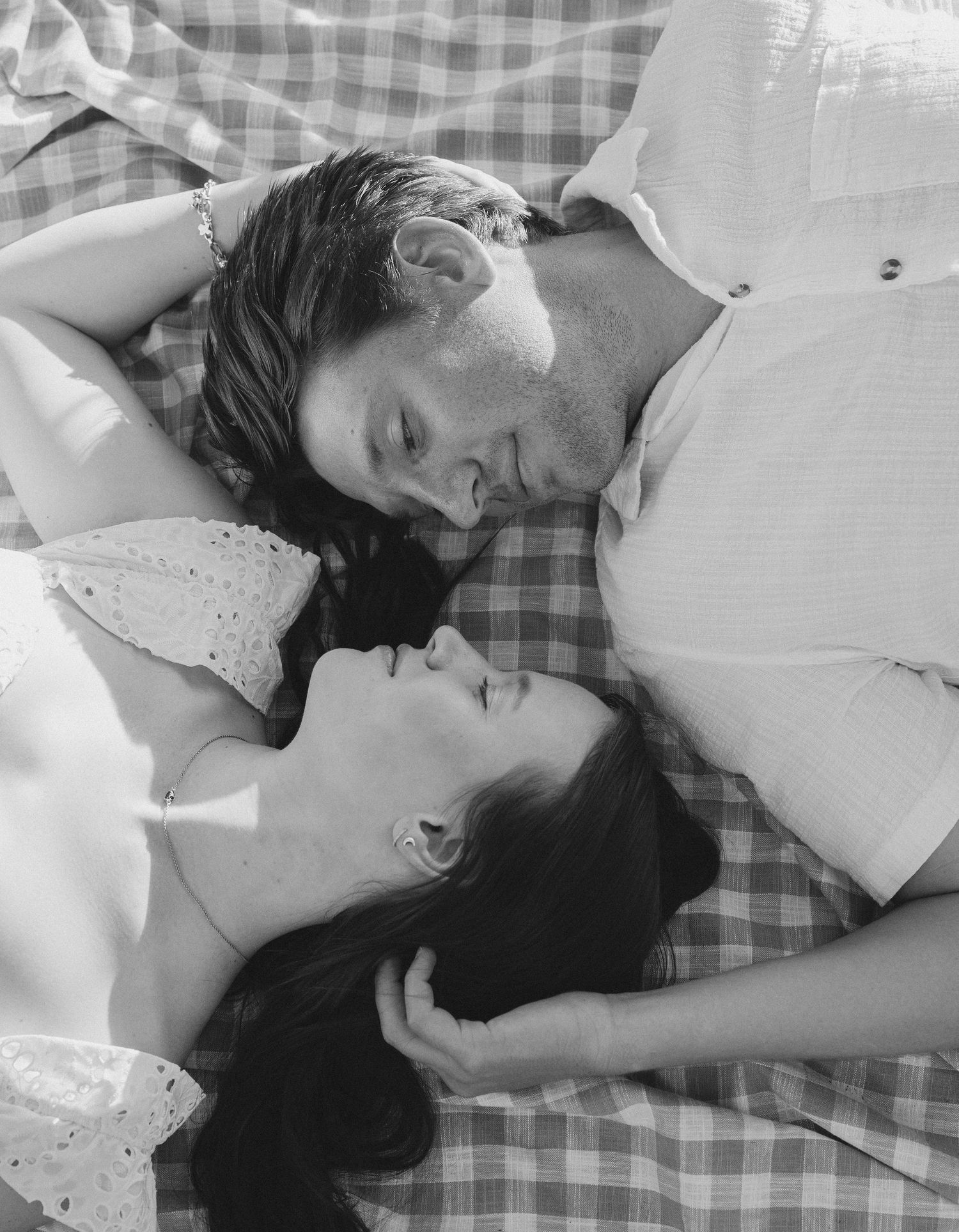 A romantic couple lying together on a gingham blanket in black and white, captured in an intimate vintage-style portrait.