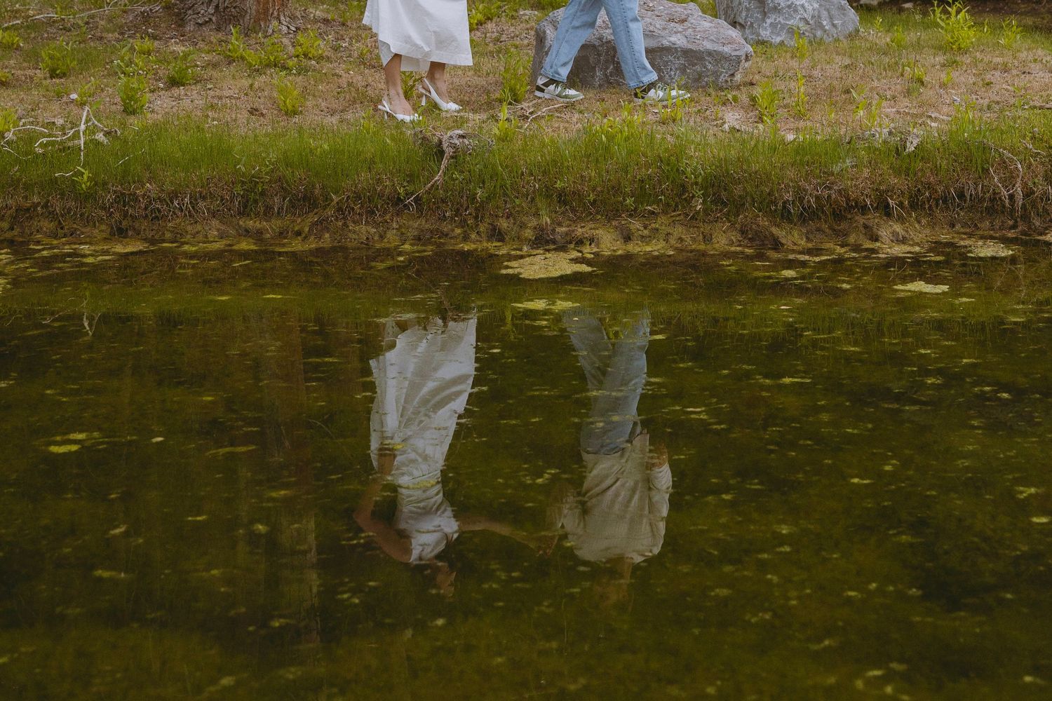 Couple in wedding attire walking by a pond with their reflection visible in the still green water below.