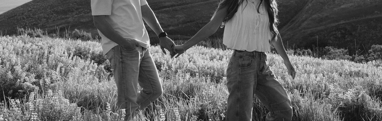 Black and white photo showing two people in casual clothes walking together through a wildflower meadow.