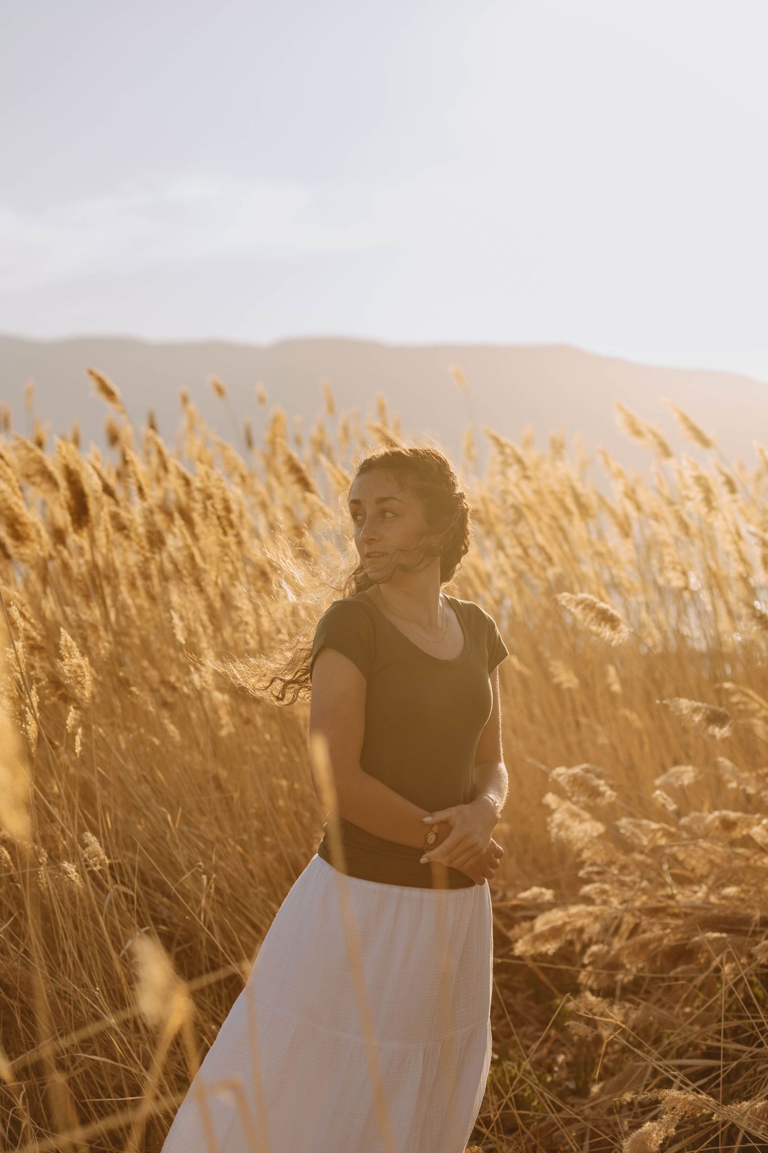 A person in a black top and white skirt stands in a golden wheat field at sunset.