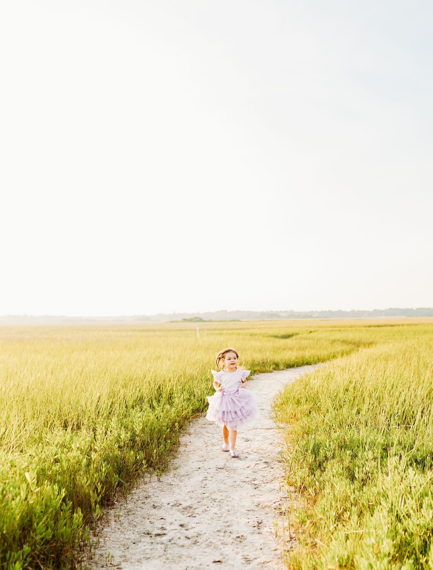 a little girl in a purple dress, runs through the marsh at sunset