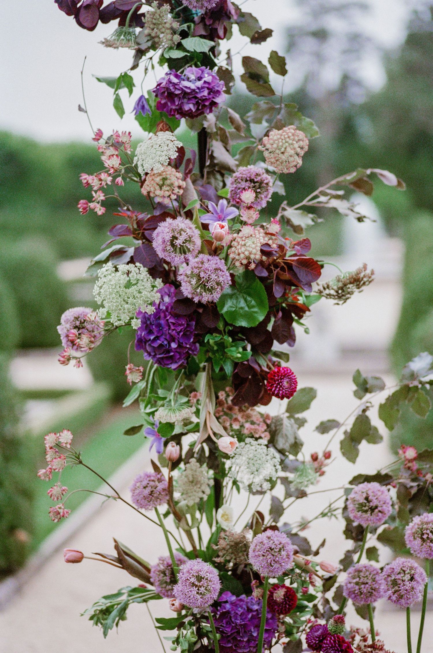 A romantic floral arrangement with purple and pink blooms including astrantia, scabiosa, and delicate foliage on display outdoors.