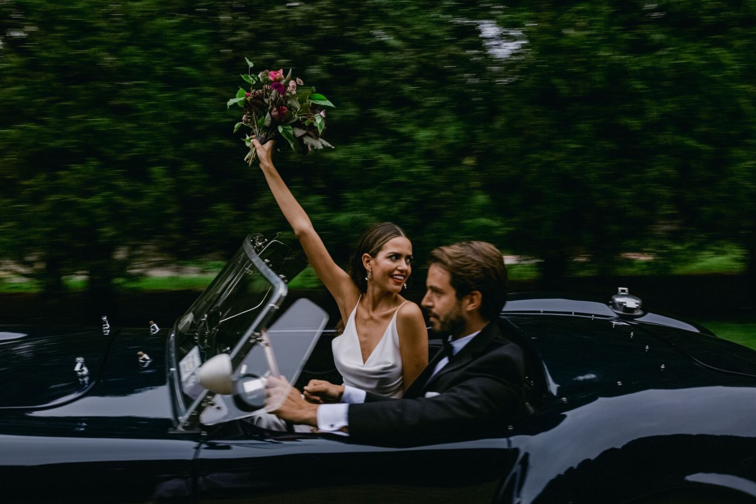 Newlyweds celebrate in a classic black convertible car while holding up a wedding bouquet during their countryside drive.