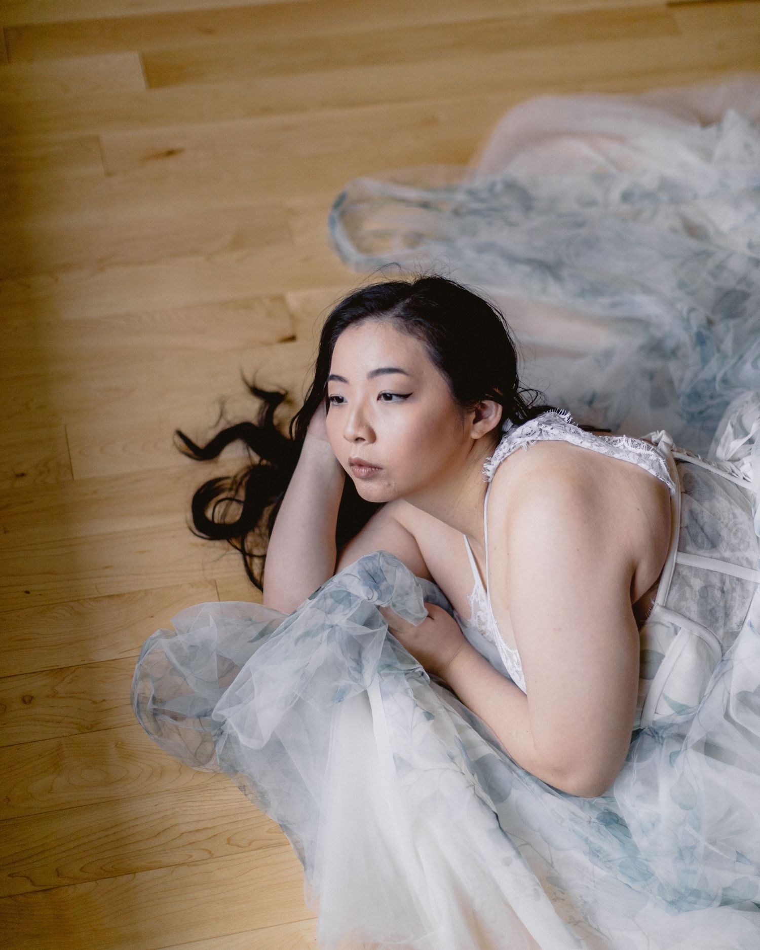 Artistic portrait of someone lying gracefully on wooden floor in flowing tulle gown with soft natural lighting.