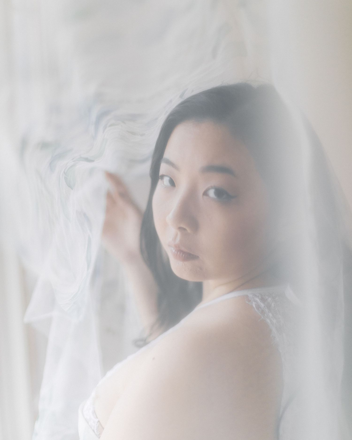 Soft ethereal portrait through delicate sheer fabric creating a dreamy atmospheric effect in muted tones.