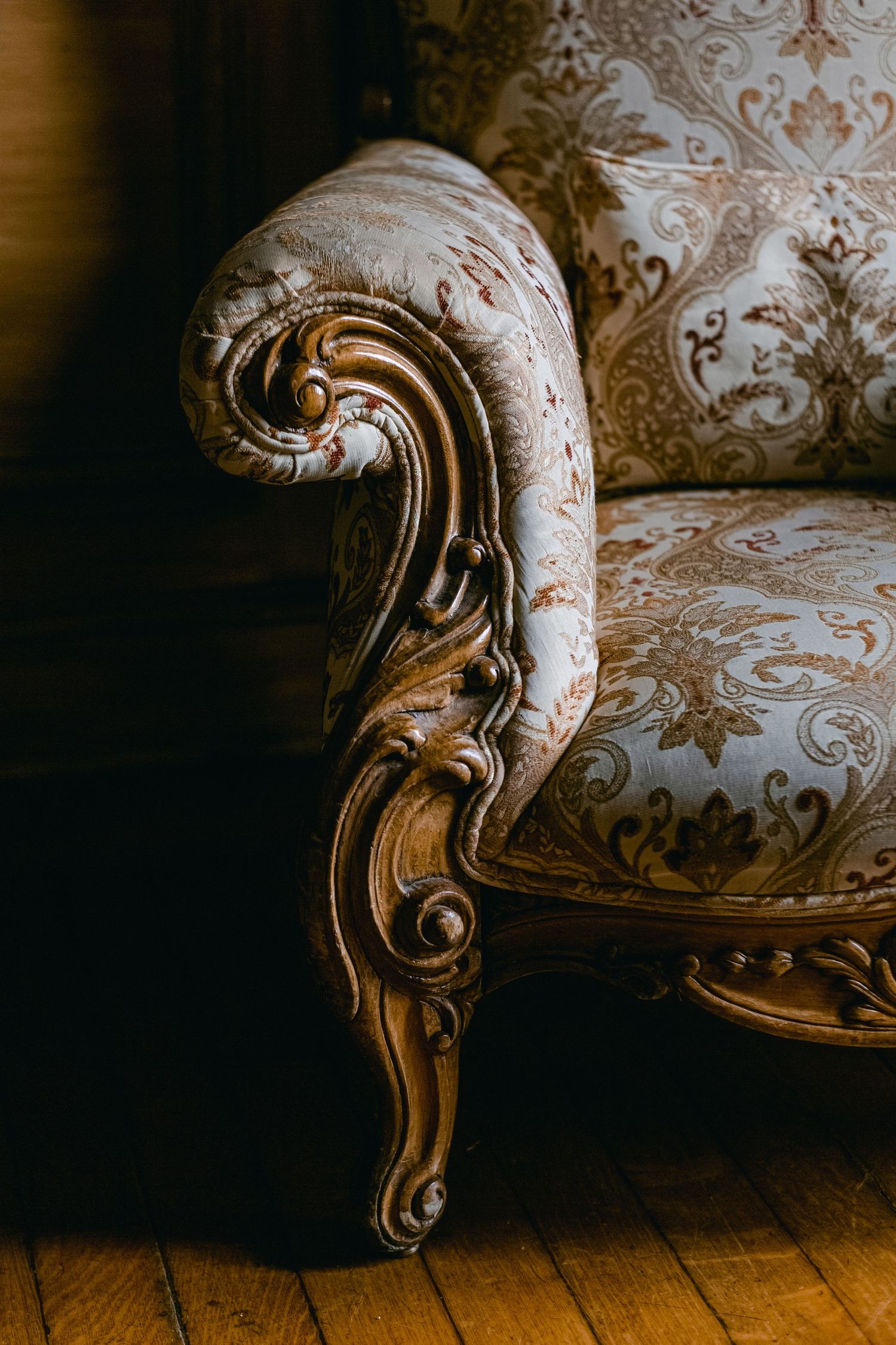 Close-up detail of an ornate vintage armchair with carved wooden scrollwork and damask upholstery on hardwood flooring.