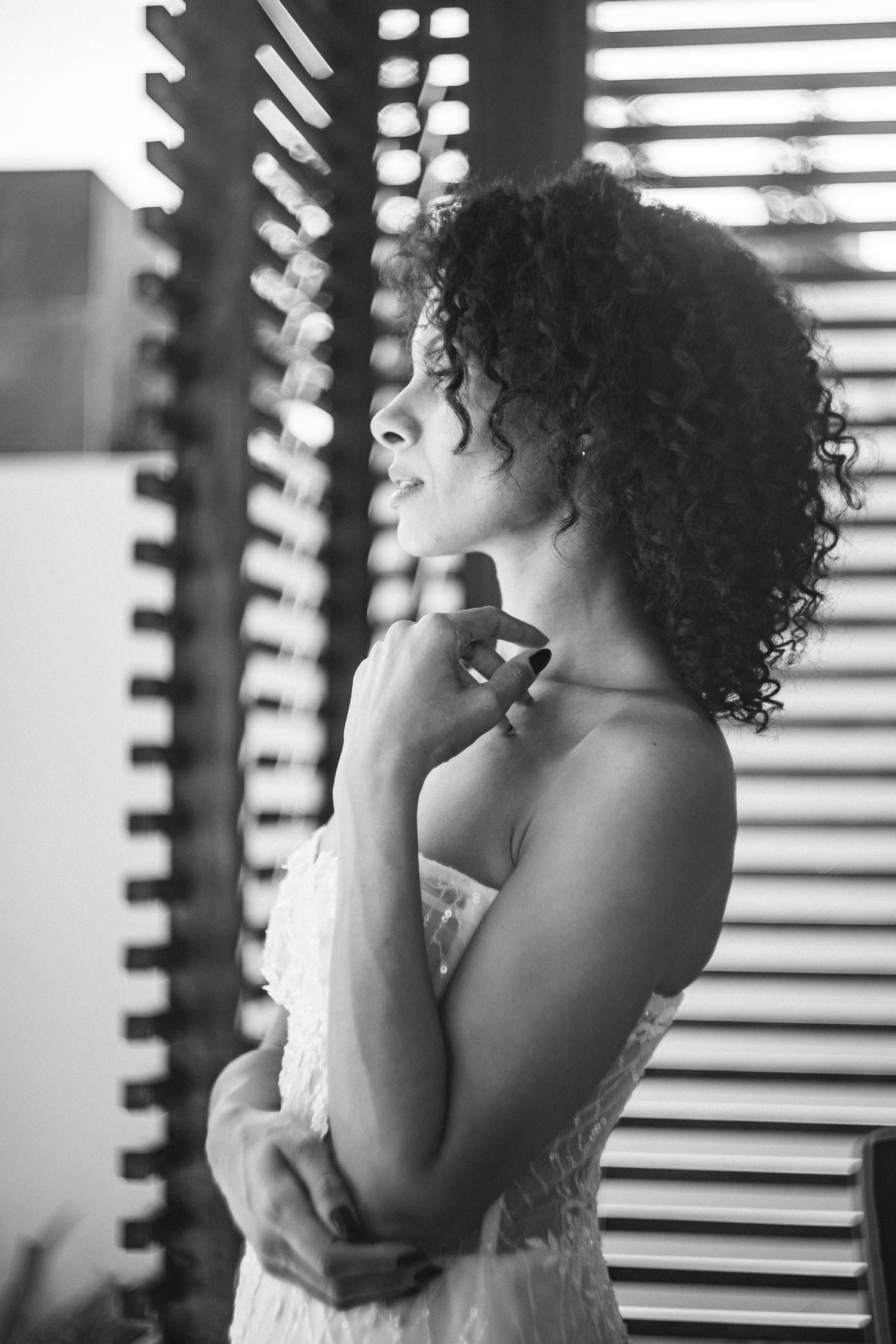 Black and white portrait of someone with curly hair in profile view by window blinds, wearing white lace.