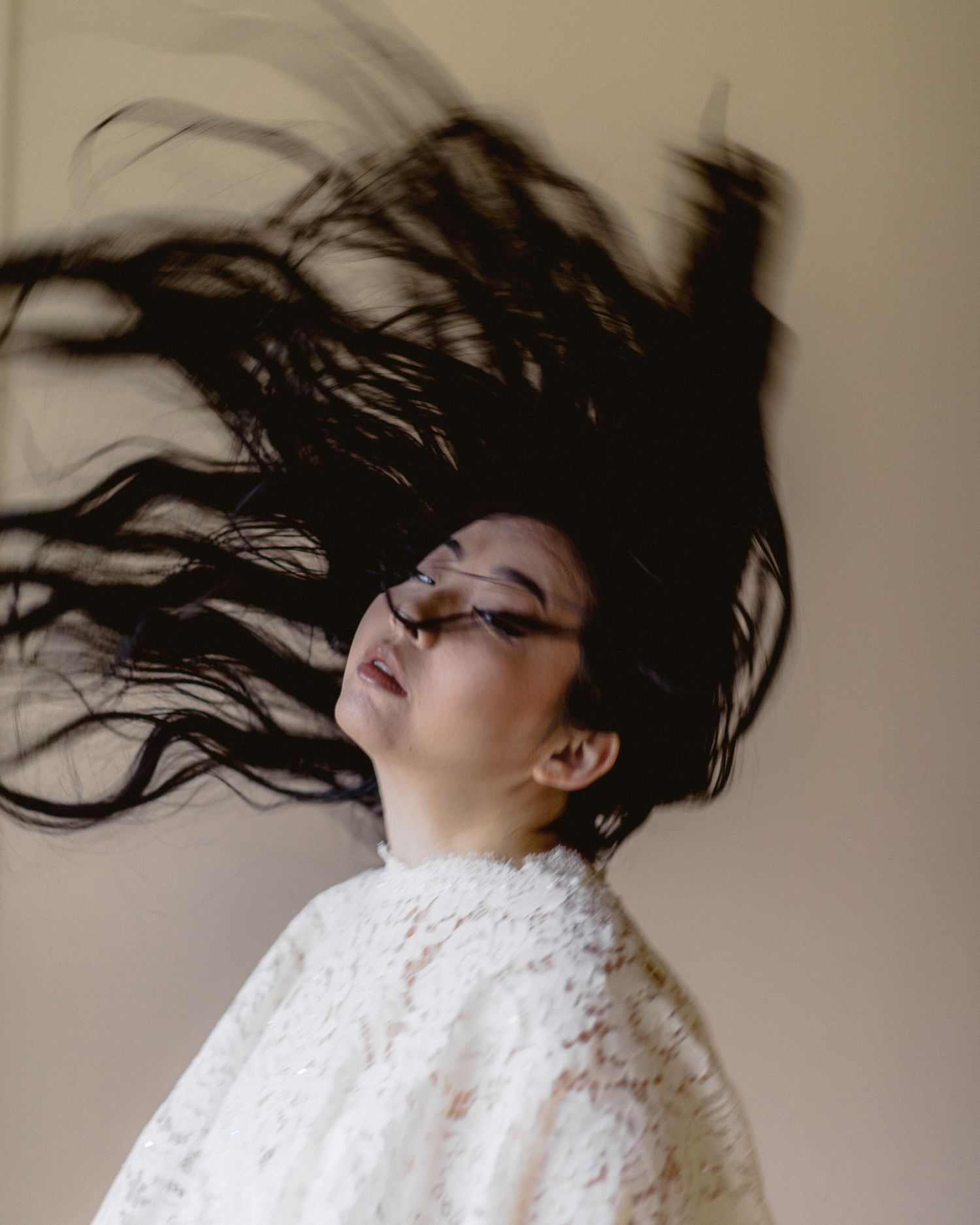 Person in white lace top with long dark hair dramatically fanned out against neutral wall.