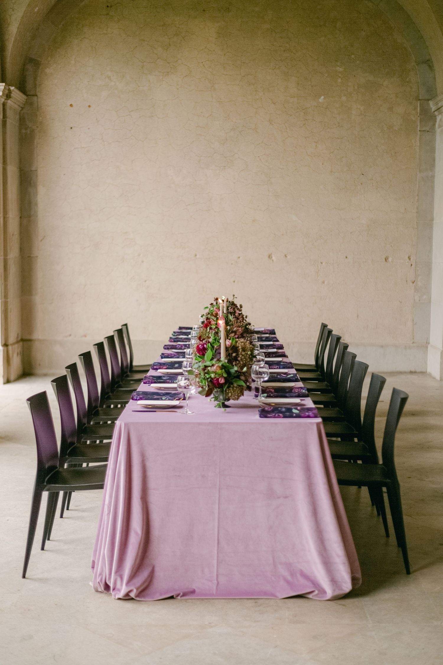Elegant dining table with purple tablecloth, black chairs and ornate floral centerpiece in an arched architectural space.