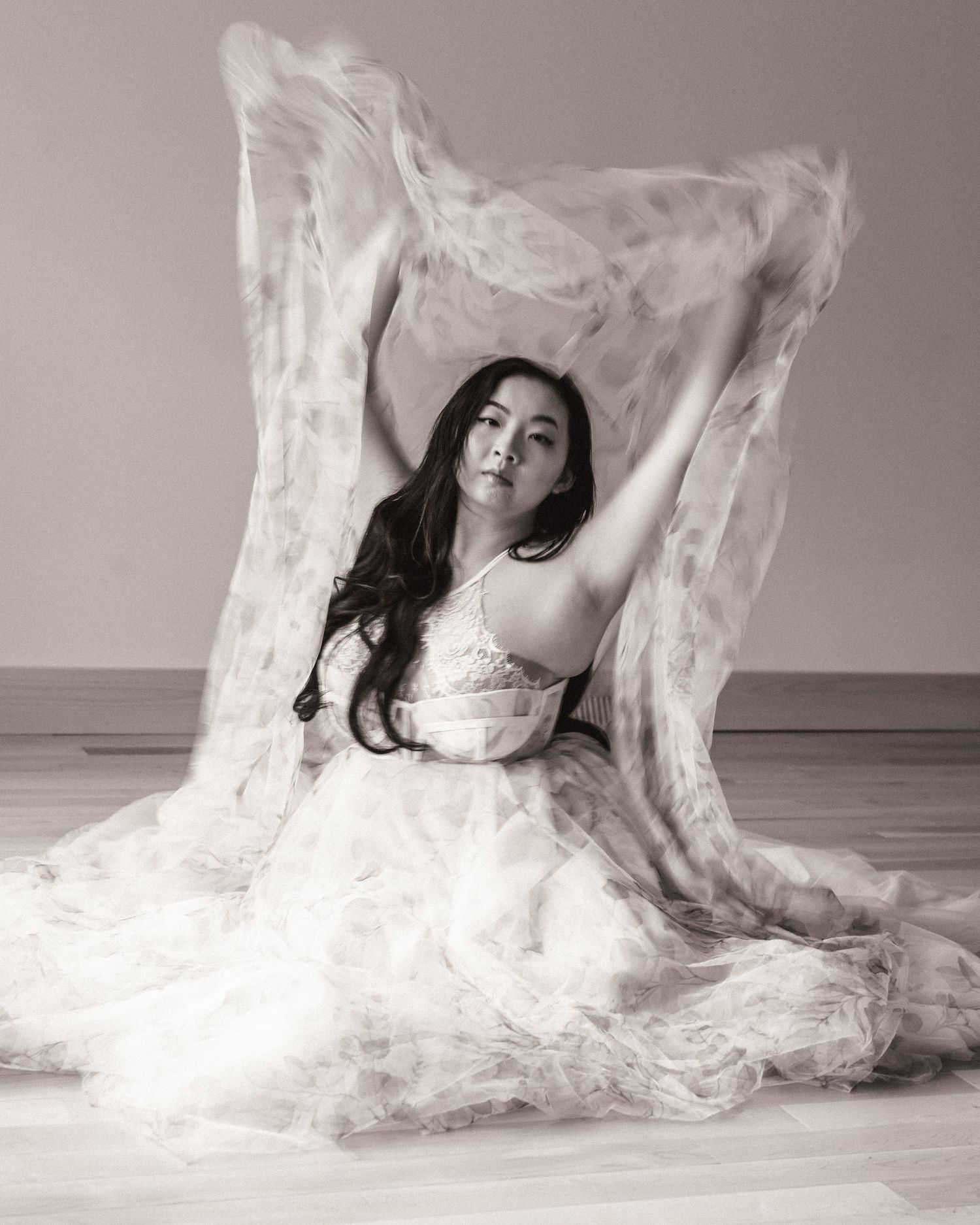 A black and white artistic portrait of a person in flowing white fabric sitting on the floor in a graceful pose.