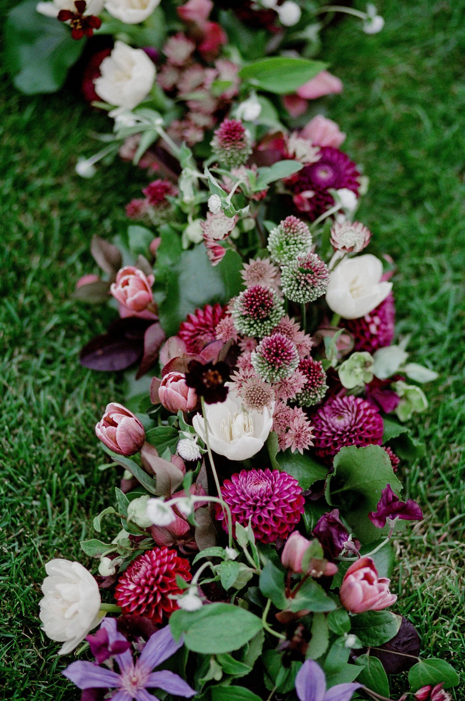 Vibrant floral arrangement with pink roses, deep burgundy dahlias, and purple blooms laying on green grass.