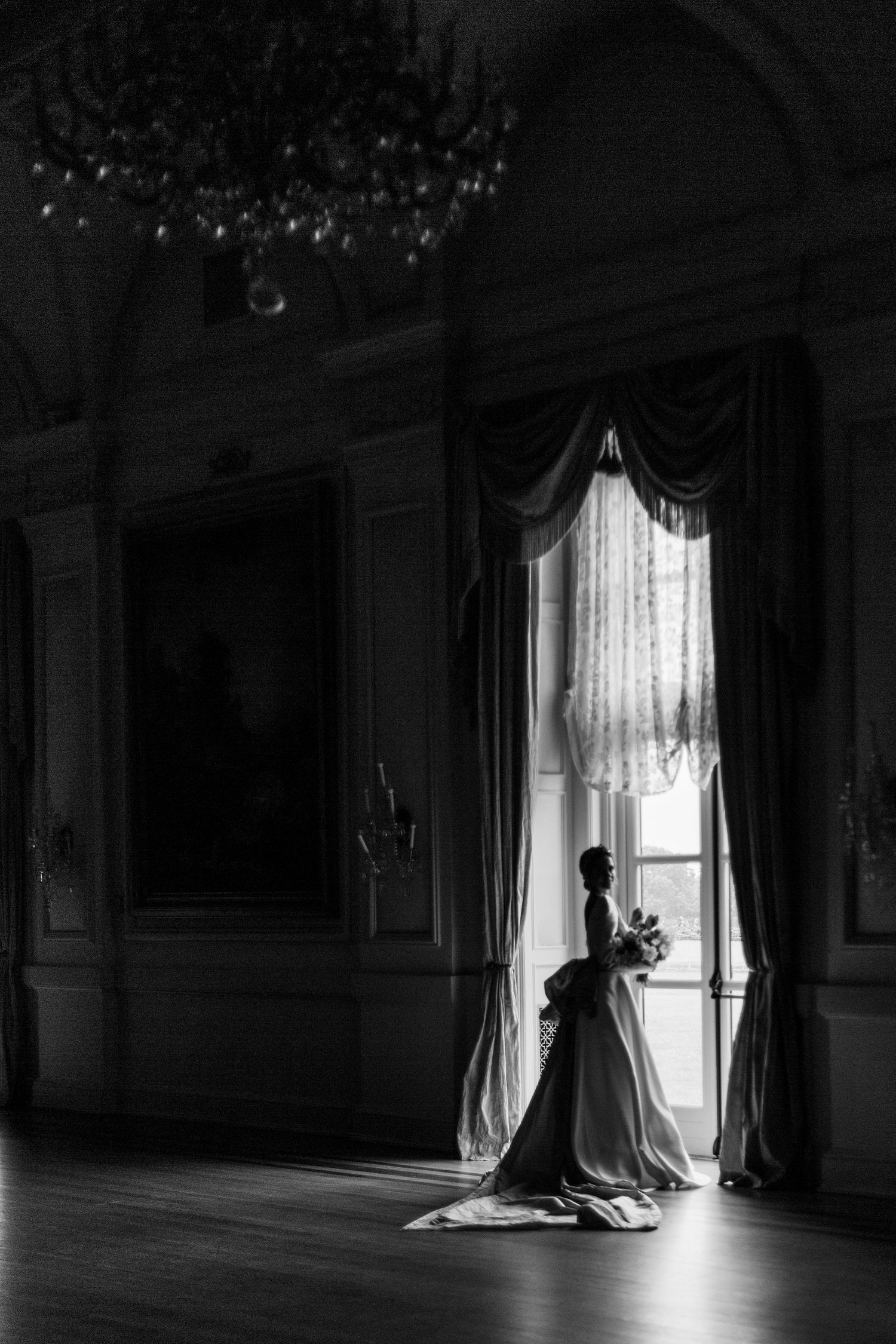 A silhouetted figure stands by an elegant window in a grand ballroom with a chandelier, creating a dramatic black and white scene.
