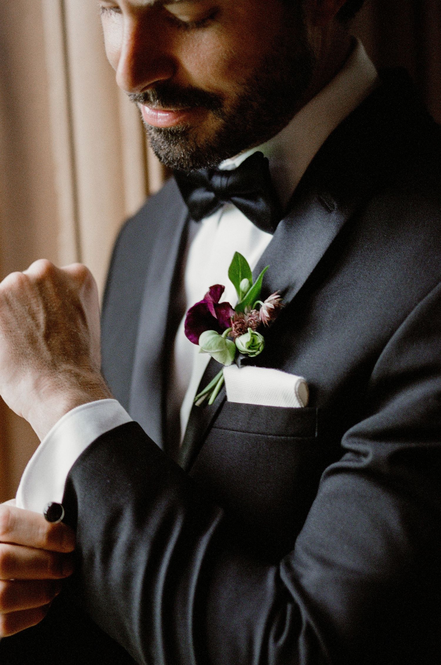 Close-up of groom in black tuxedo and bowtie adjusting cufflink, with deep purple boutonniere on lapel.
