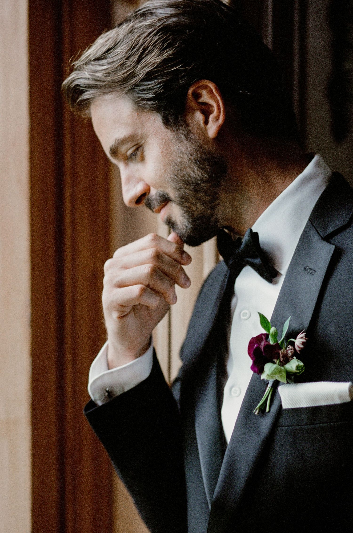 Side profile of groom in classic black tuxedo with bowtie and floral boutonniere laughing during wedding preparations.