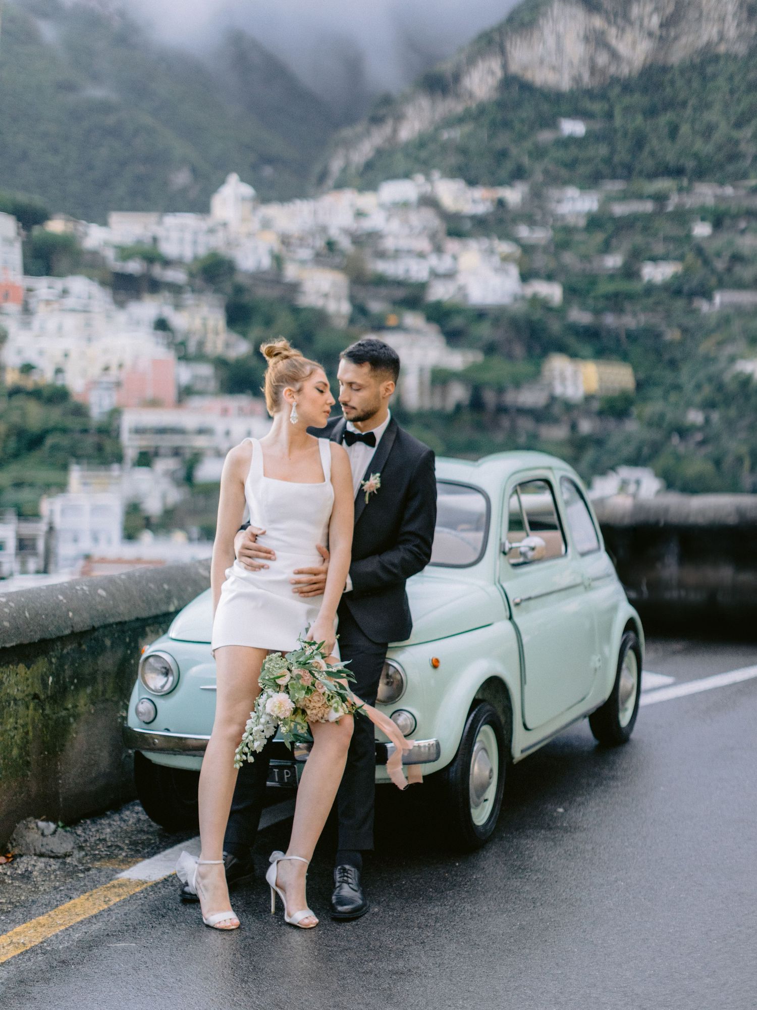 A vintage Fiat 500 parked on a cliffside road in Positano, Italy with coastal buildings cascading down the mountainside.