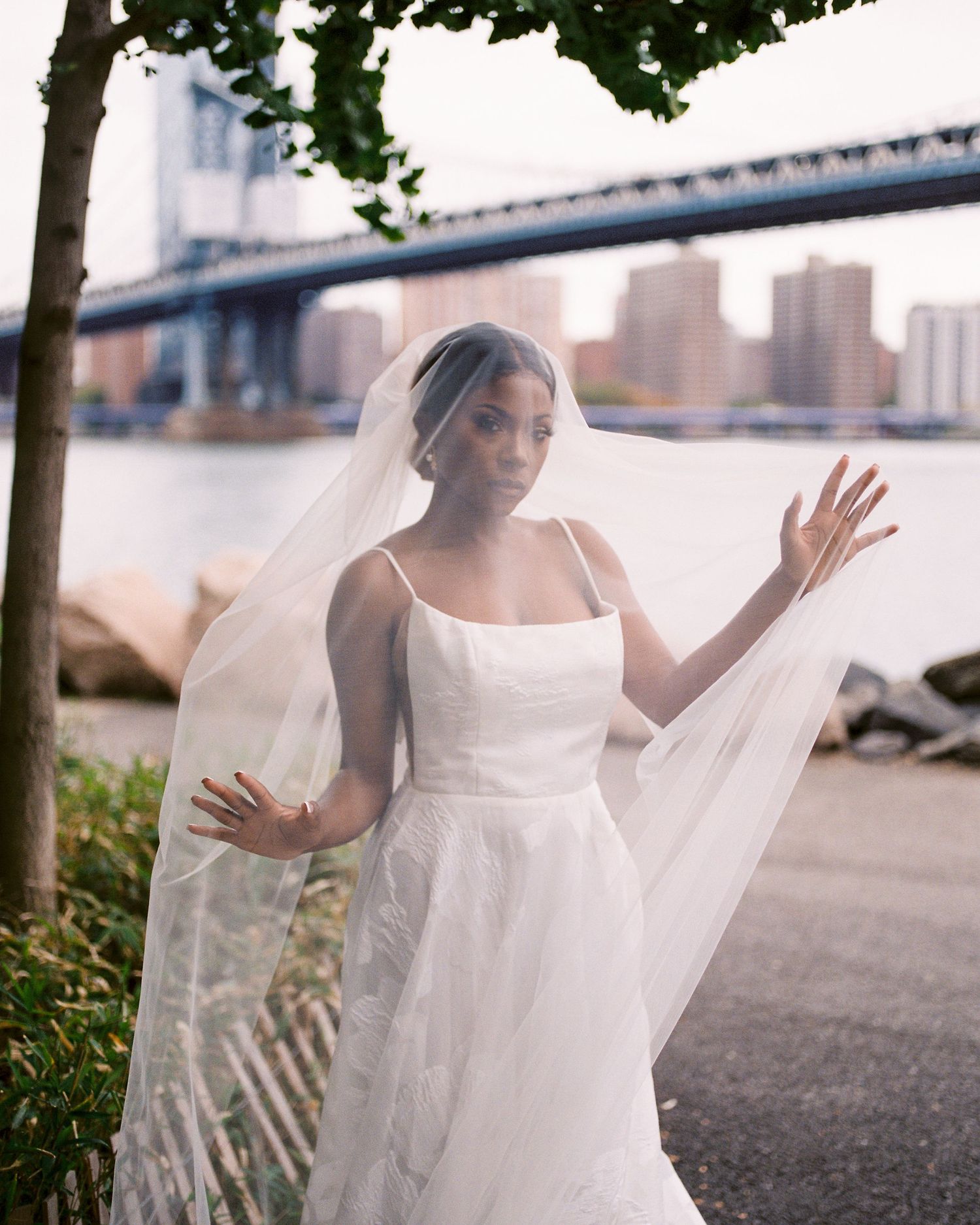 A bride in a classic white wedding dress and flowing veil poses gracefully by the Manhattan Bridge waterfront in Brooklyn.