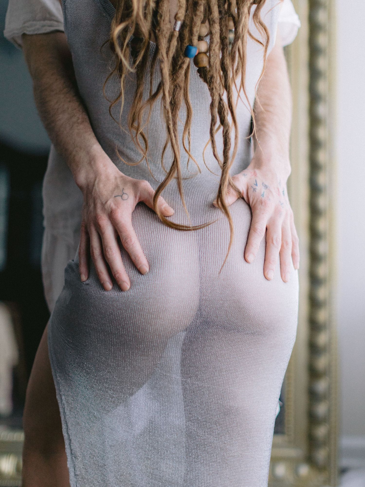 intimate photo of long-haired woman wearing sheer dress poses with partner's hands on butt with ornate mirror frame in New Hope studio.