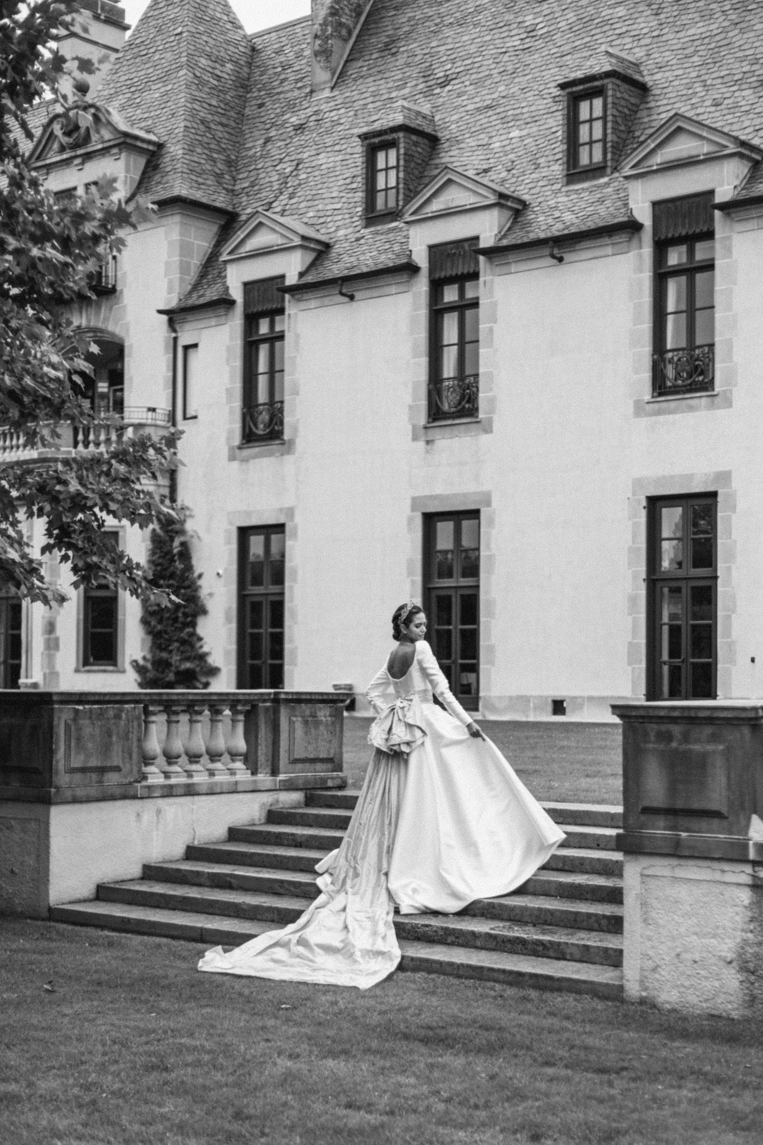 A vintage black and white photo shows an elegant wedding gown with long train on the grand steps of a historic chateau.