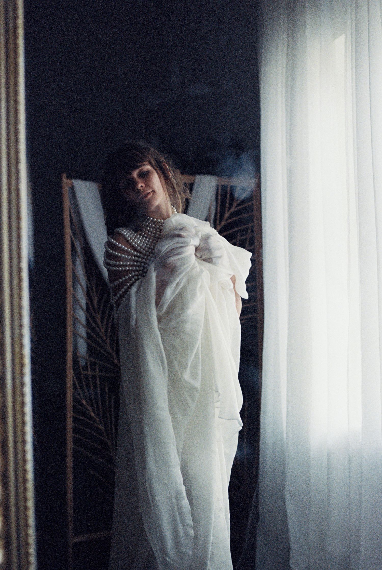 Moody artistic portrait in dark room with white fabric and sheer curtains creating ethereal atmosphere.