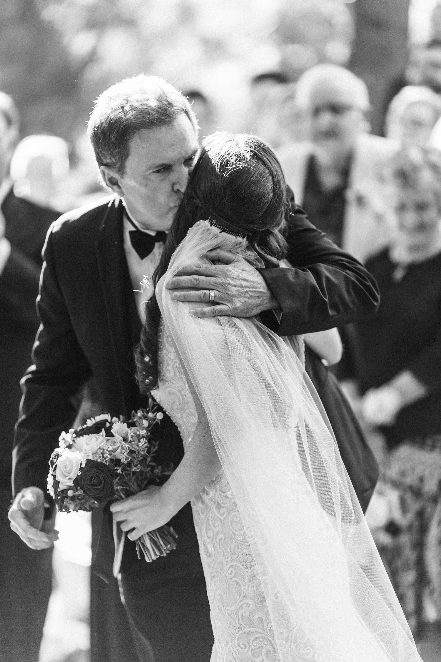 Black and white emotional moment of dad and bride during outdoor wedding ceremony with guests in background.