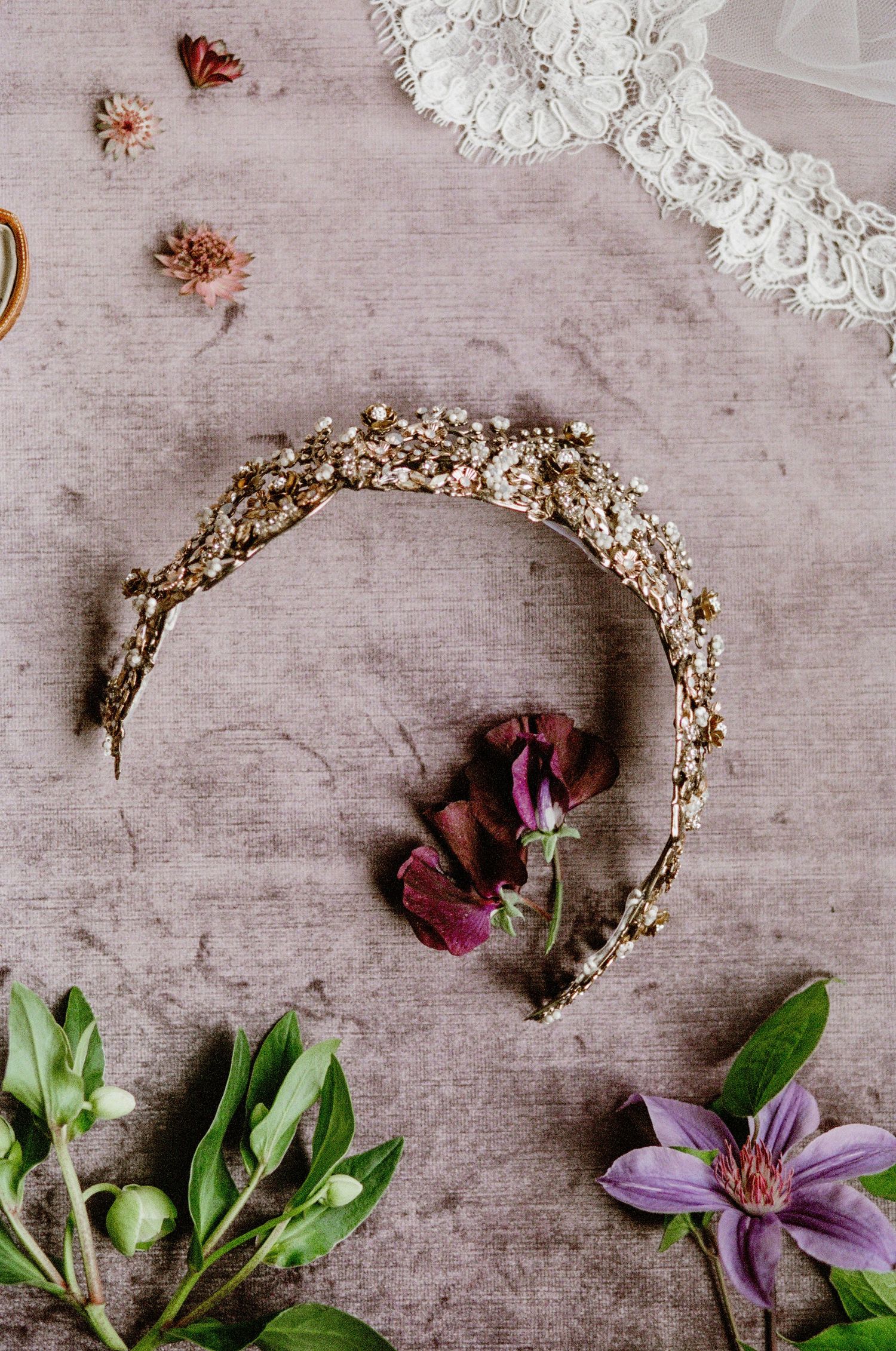 Vintage lace trim and ornate golden headpiece arranged with dried roses and purple clematis on textured fabric background.