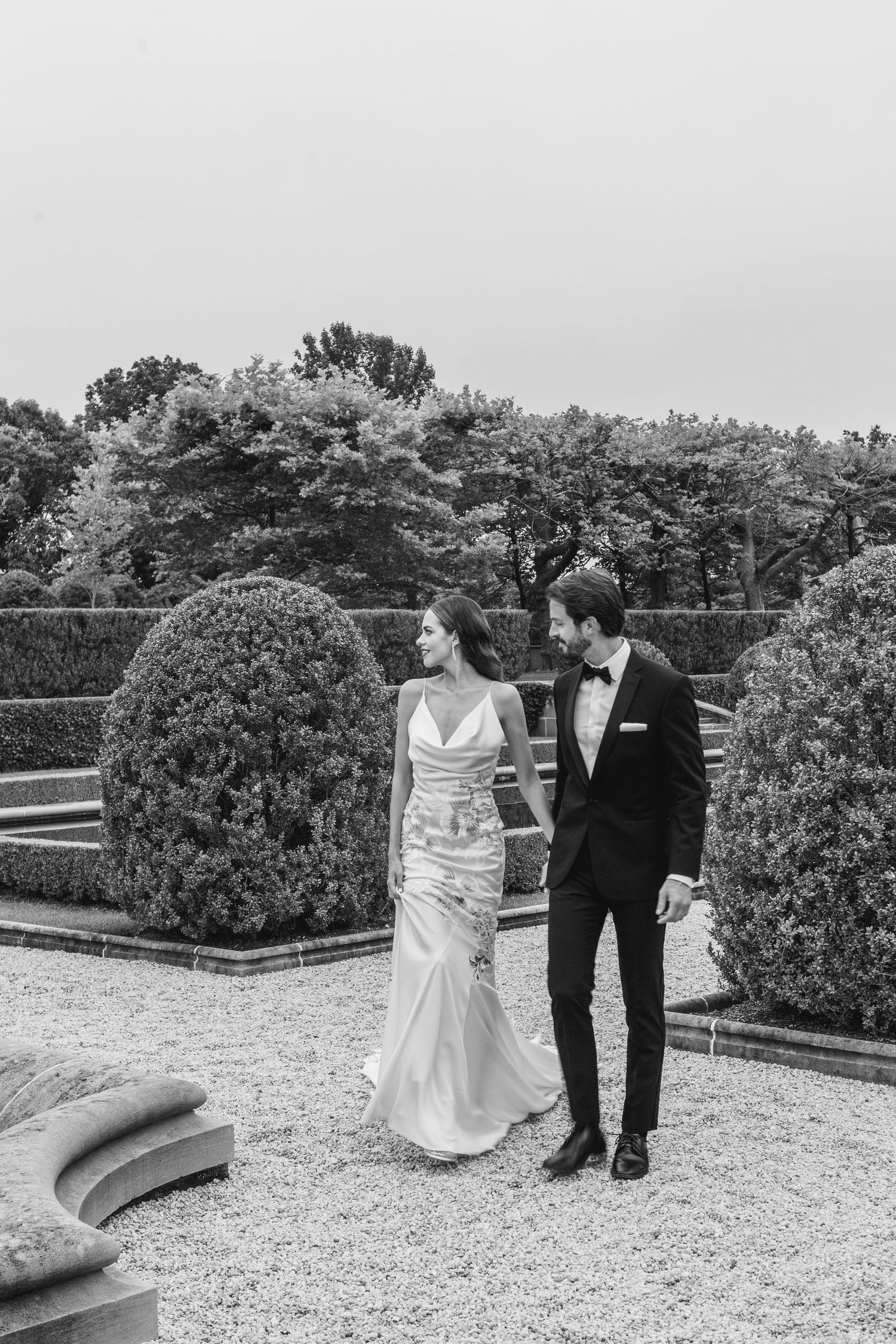 A bride and groom share a romantic moment on a gravel path in a formal garden with manicured shrubs and trees.