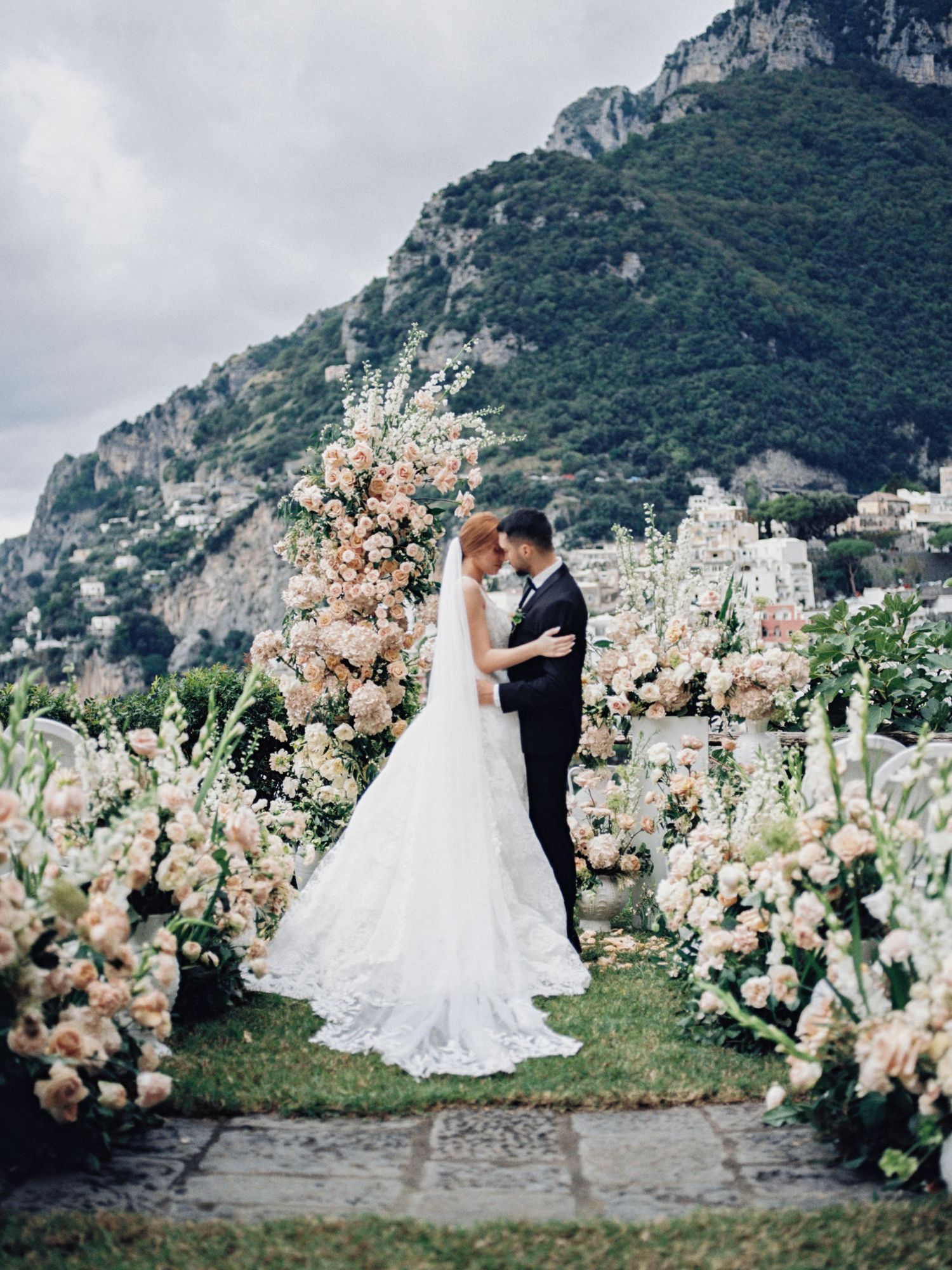 Wedding couple embraces in romantic Italian garden with mountain backdrop and luxurious floral arrangements in Positano. Monique Lhuillier